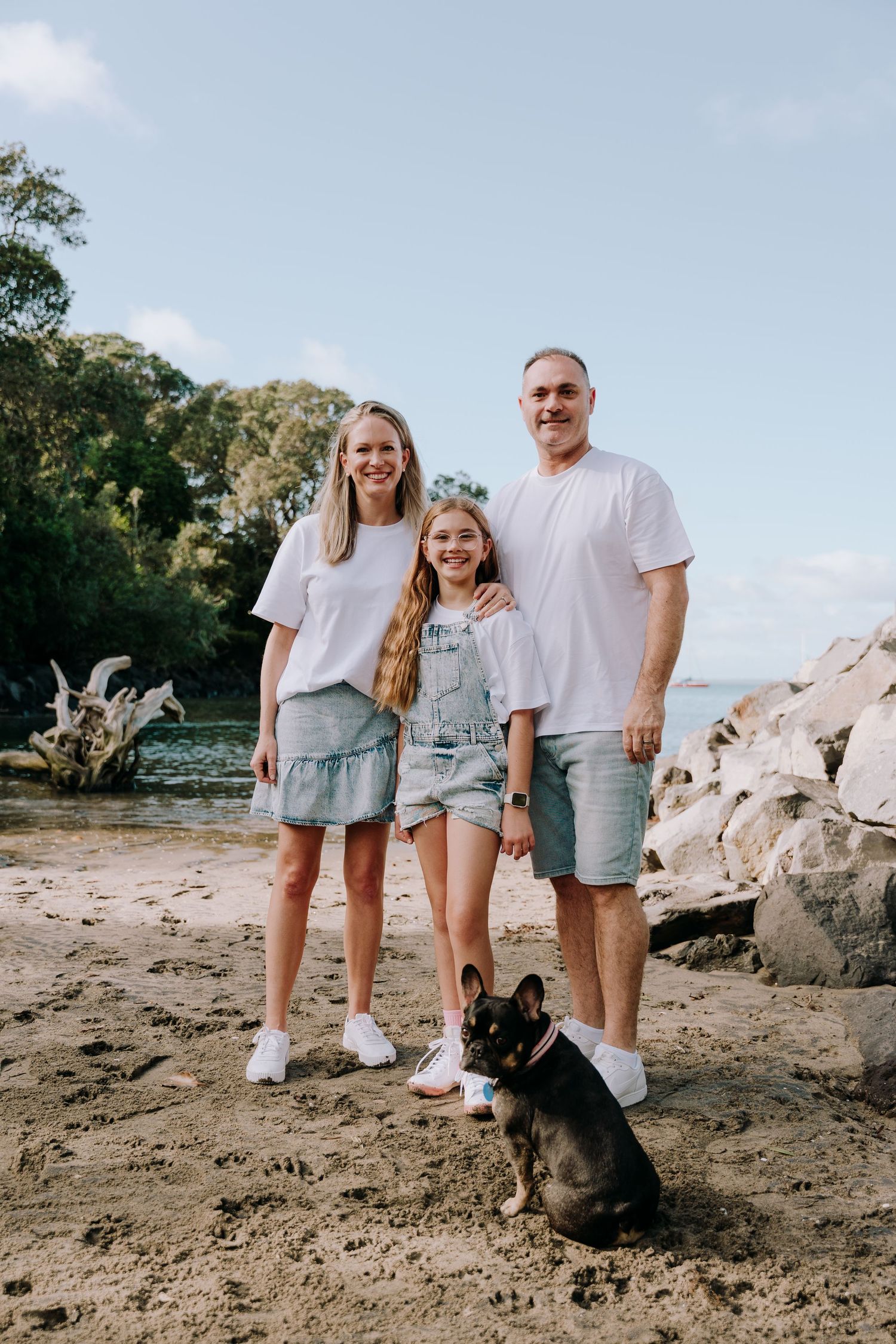 Family in coordinated casual outfits posing with their French Bulldog on a rocky beach.