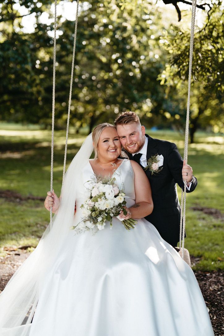 Newlyweds share romantic moments on white wooden swings in an outdoor garden setting.