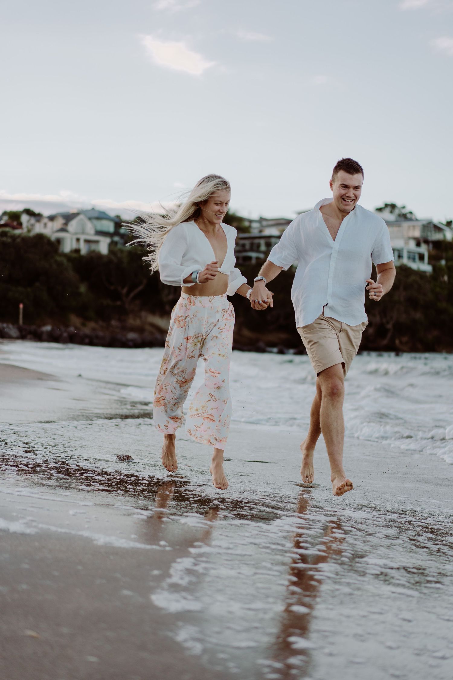 A couple playfully splashes and runs through shallow ocean water at sunset on a sandy beach.
