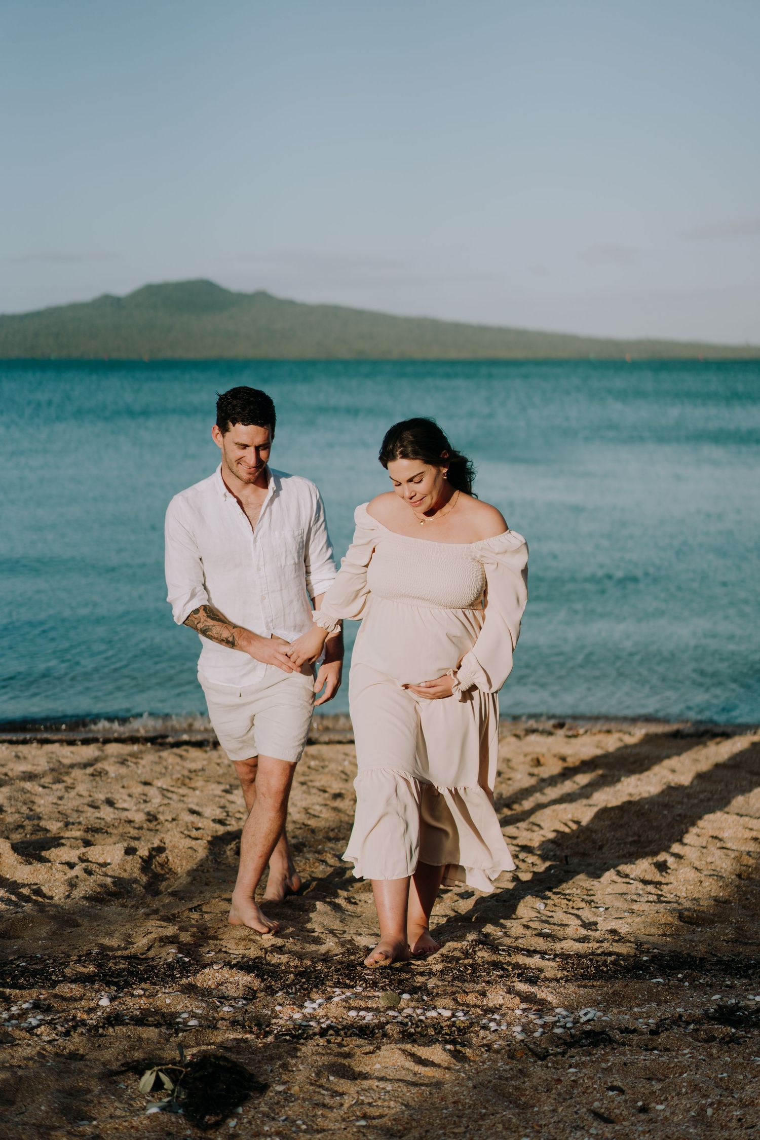 A couple walks along a scenic beach with turquoise water and mountains in the background during a maternity photoshoot.