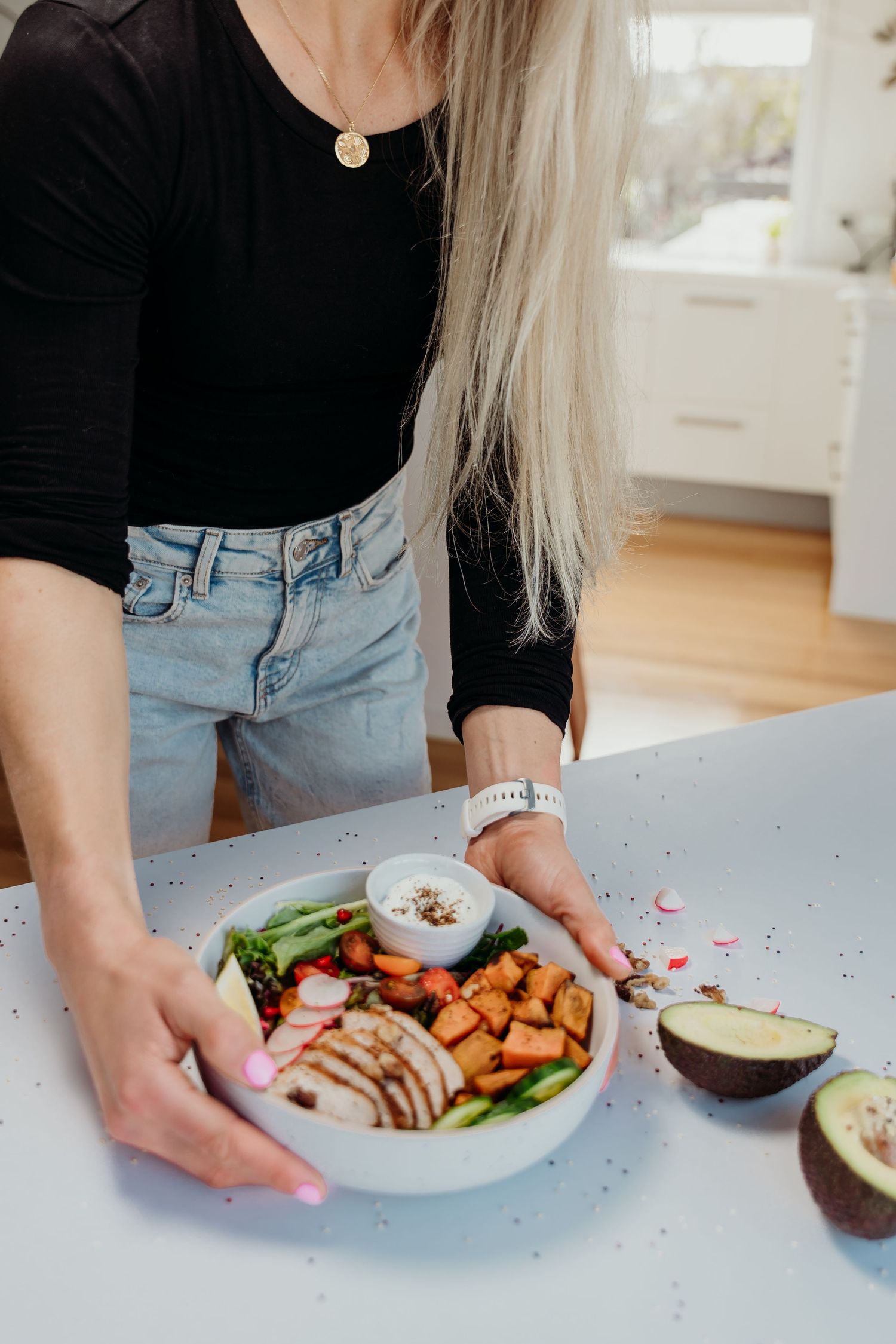 Woman placing a healthy grain bowl with chicken, roasted vegetables, and avocado on a light blue kitchen counter.
