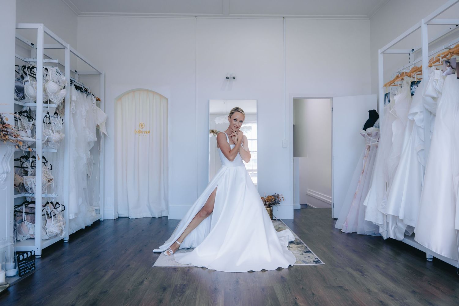 Bride posing in a white wedding gown with a high slit inside an elegant bridal boutique with dresses on display.