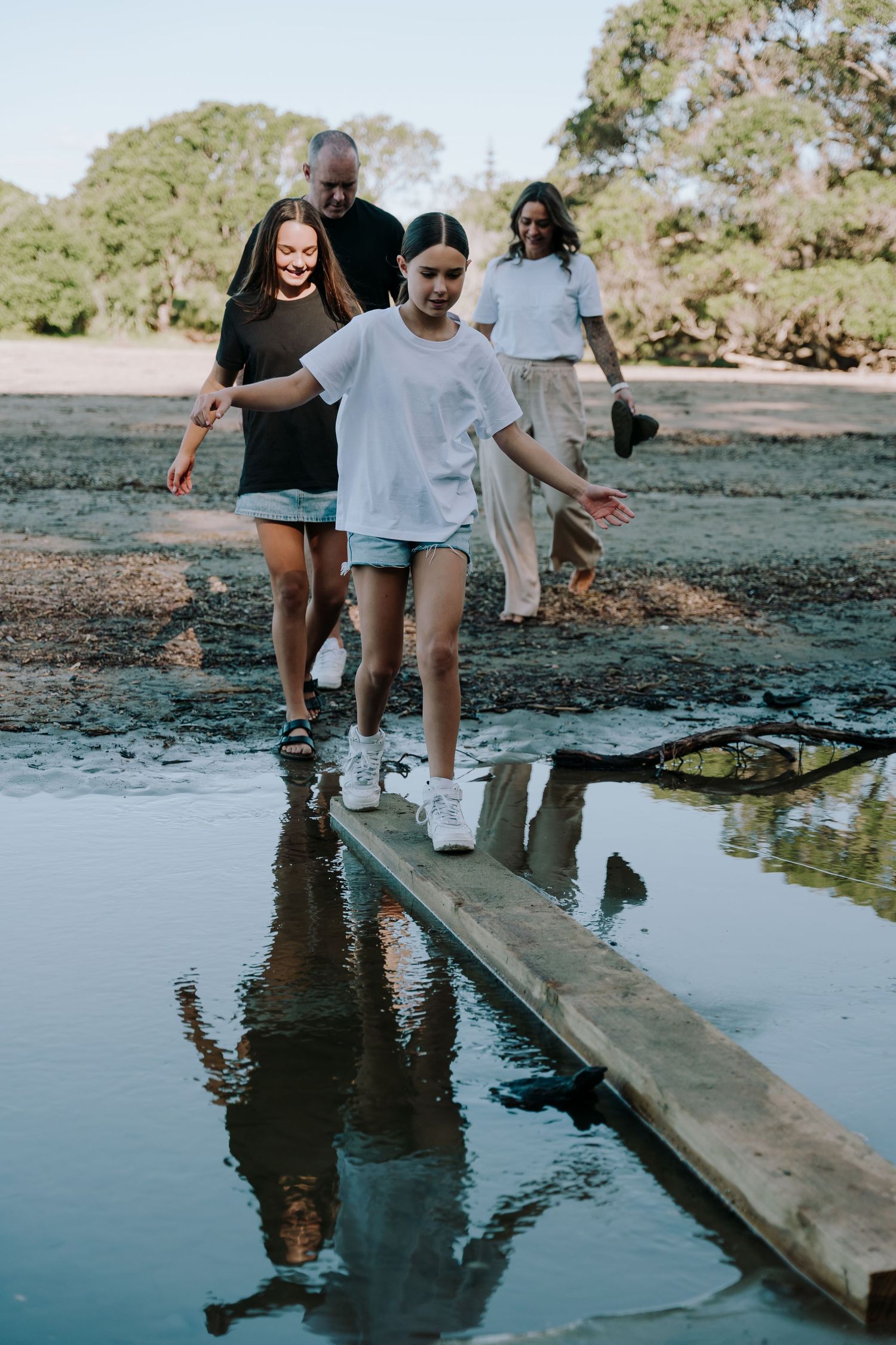 A group walks carefully across a wooden plank over still water, creating reflections in the calm surface below.