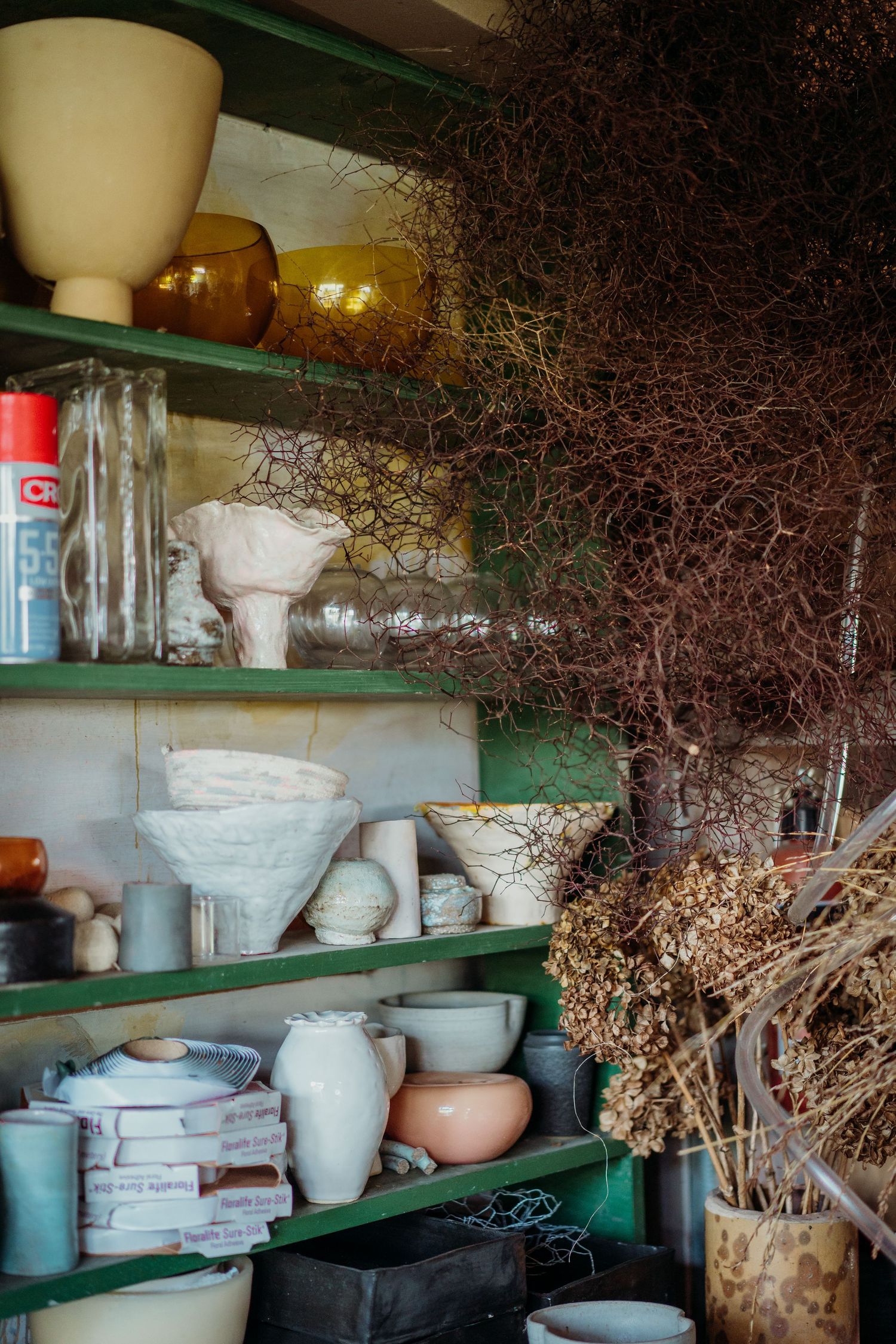 Green shelves display white ceramic vases, pottery, and dried branches in a rustic workshop setting.