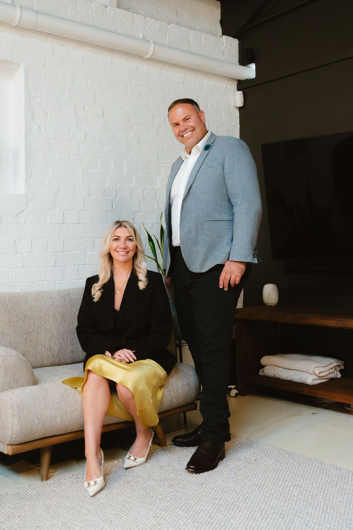 A smiling woman in black blazer and gold skirt sits on a sofa beside a tall man in grey blazer in a modern interior.