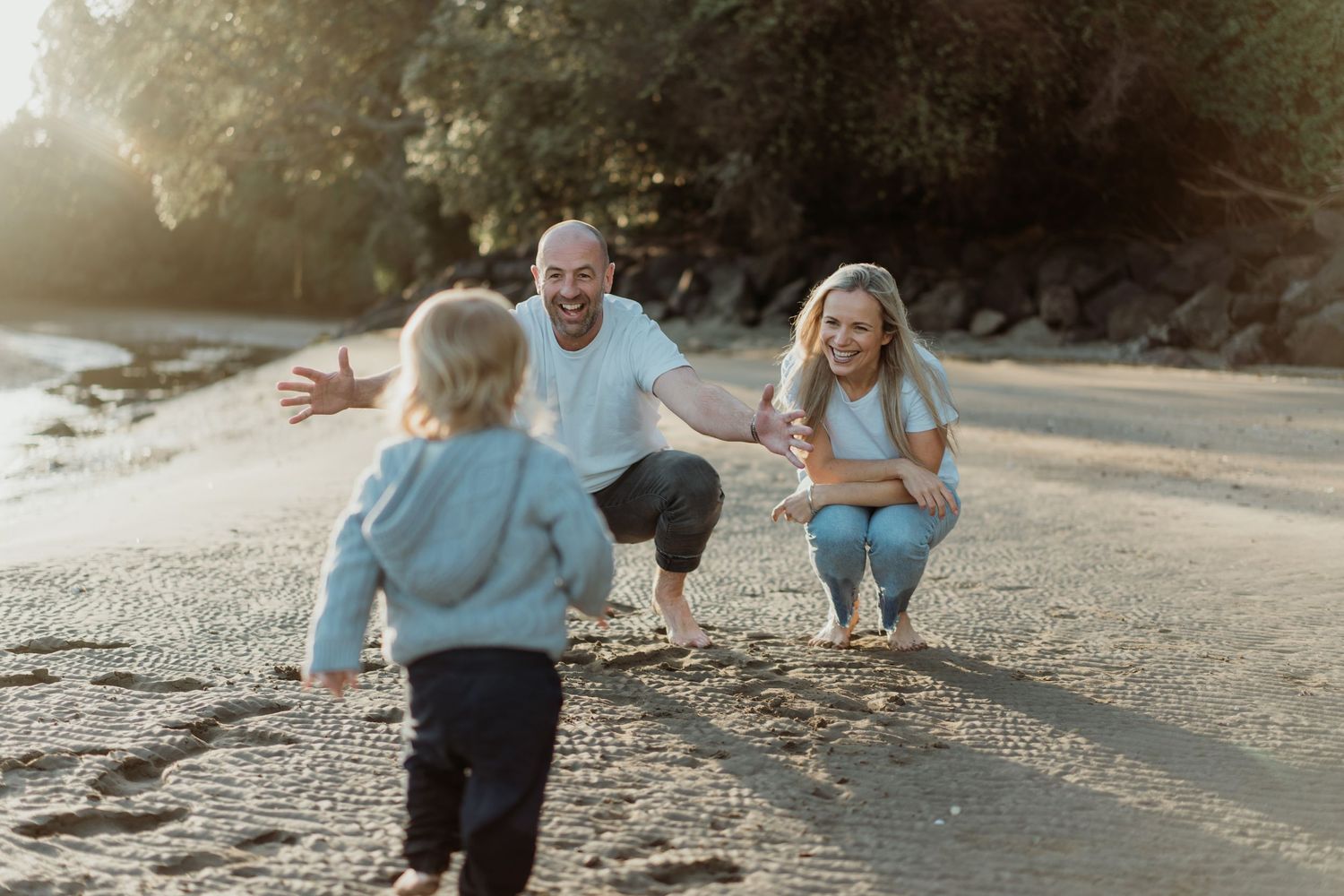 Family enjoying a sunny beach day, with child running toward parents who crouch with open arms on sandy shore.