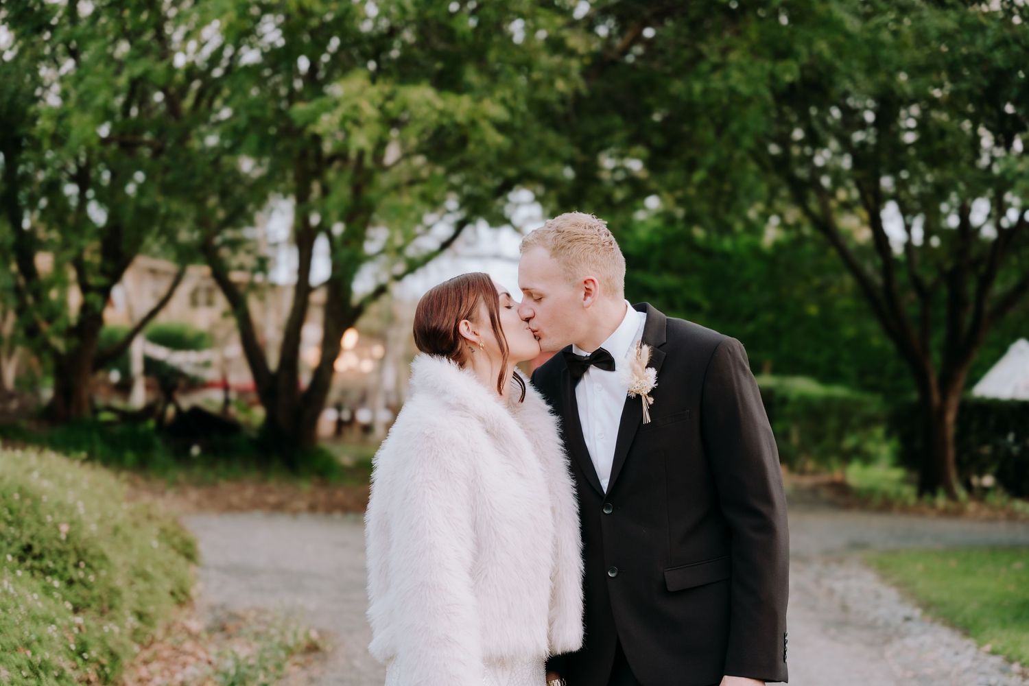 A bride in a white fur coat and groom in a black tuxedo share a kiss outdoors at their wedding.