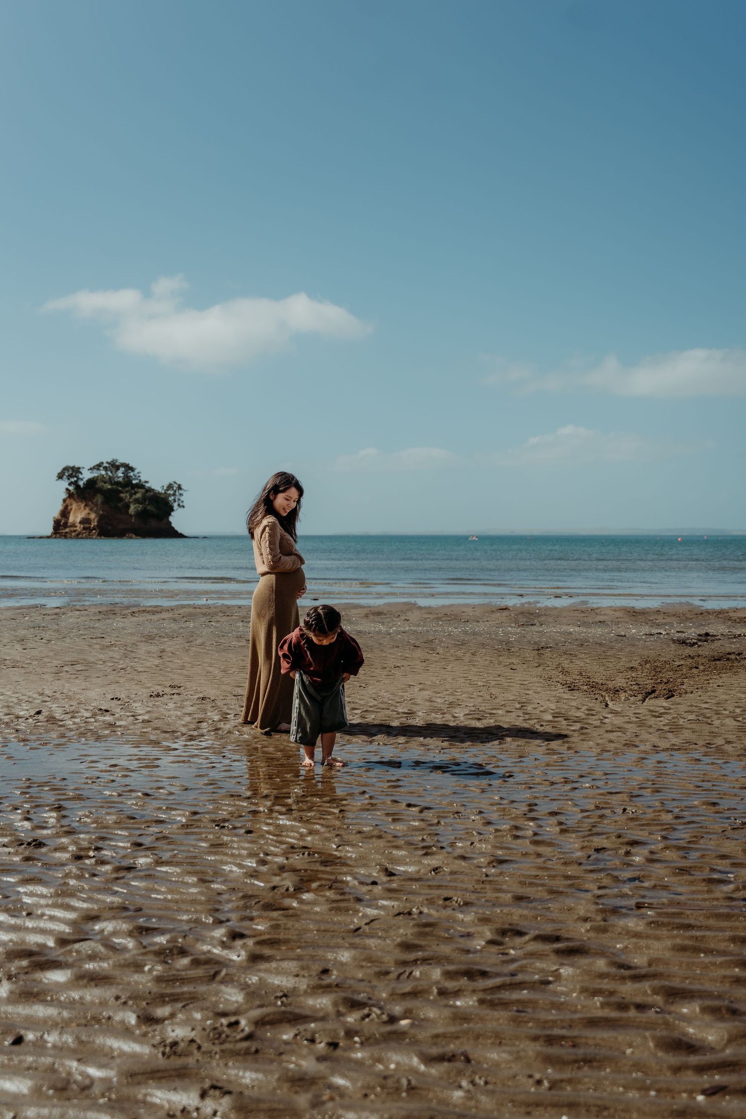 A woman in a brown dress stands on a sandy beach with a young child, ocean and small island visible behind them.