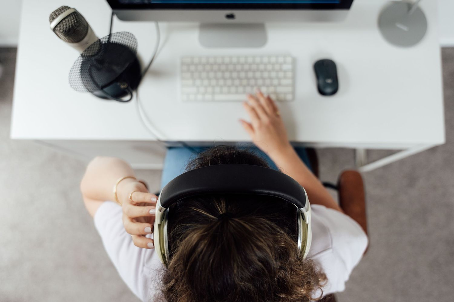Overhead view of a person wearing headphones working at a white desk with a computer, keyboard, mouse, and microphone.
