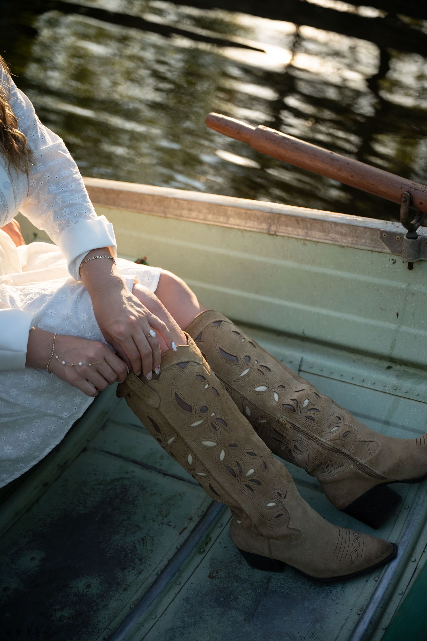 Woman in white dress wearing tan knee-high cowboy boots with cutout details, sitting in a wooden boat.