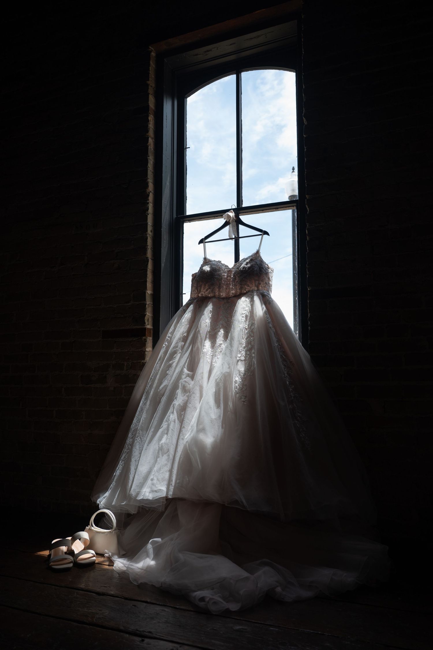 Elegant ballgown wedding dress hanging in dark window with bridal shoes on wooden floor below.