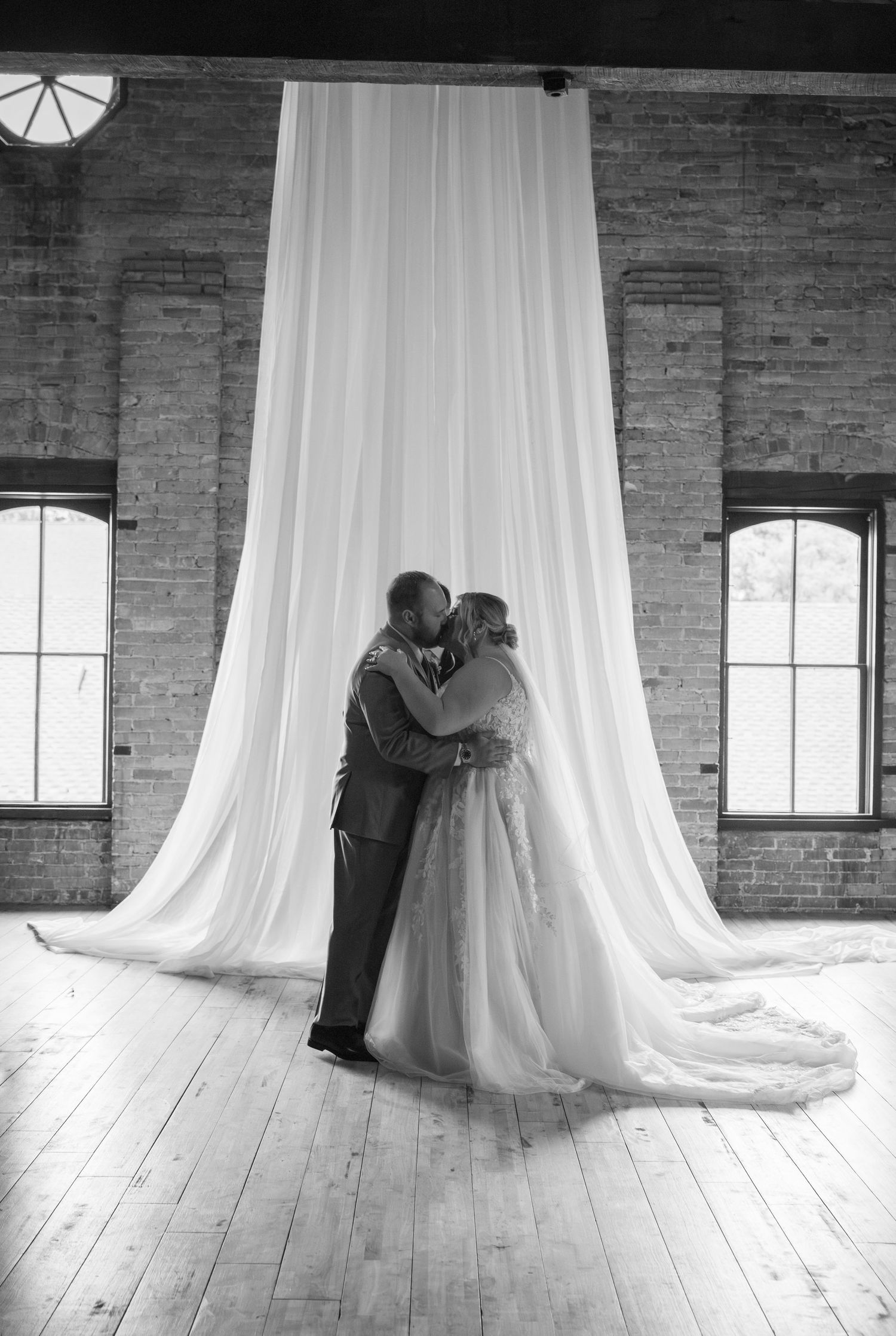 Black and white photo of bride and groom sharing first kiss at altar with flowing white drape backdrop.