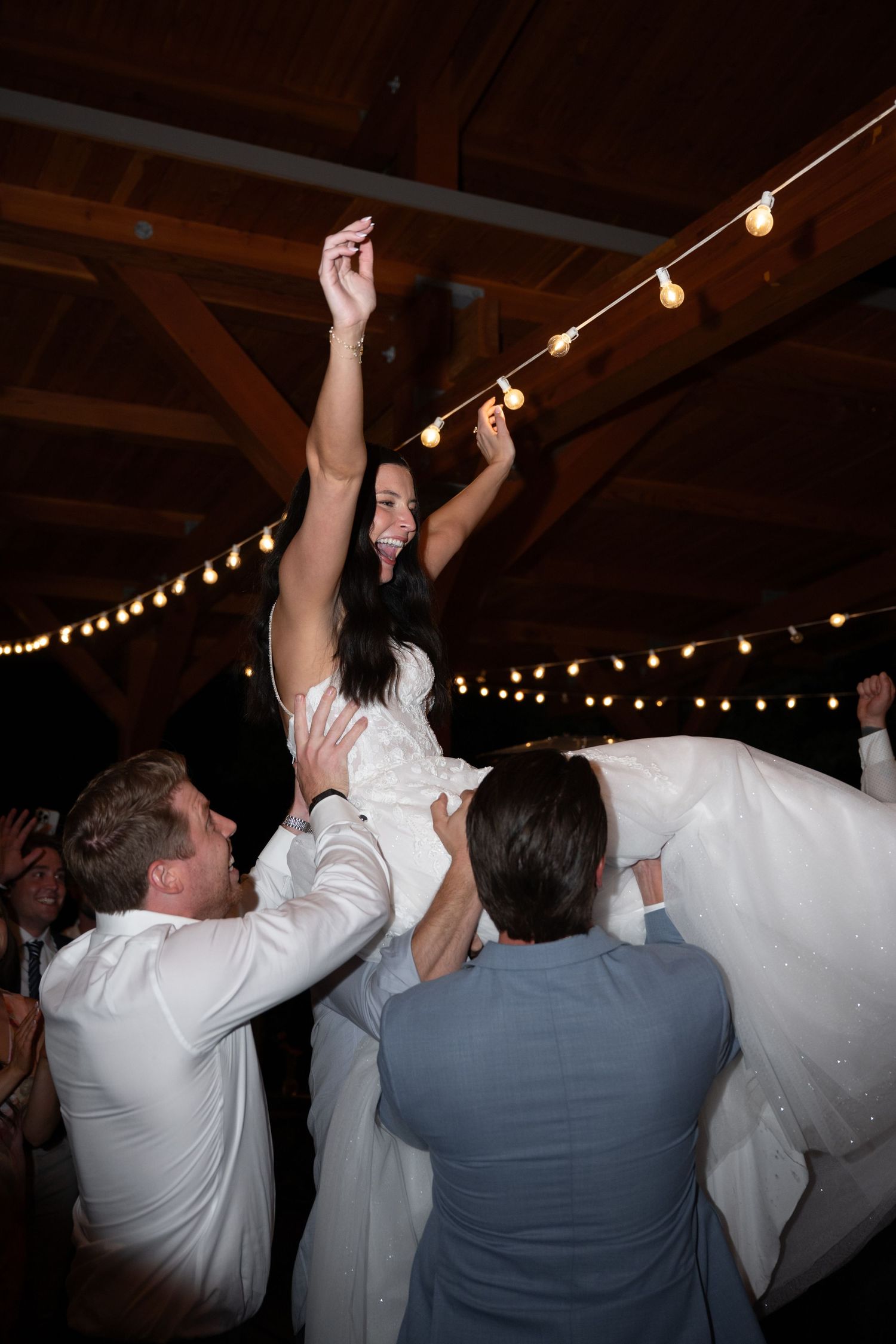Bride being lifted by groomsmen on the dance floor under string lights at a festive wedding reception.