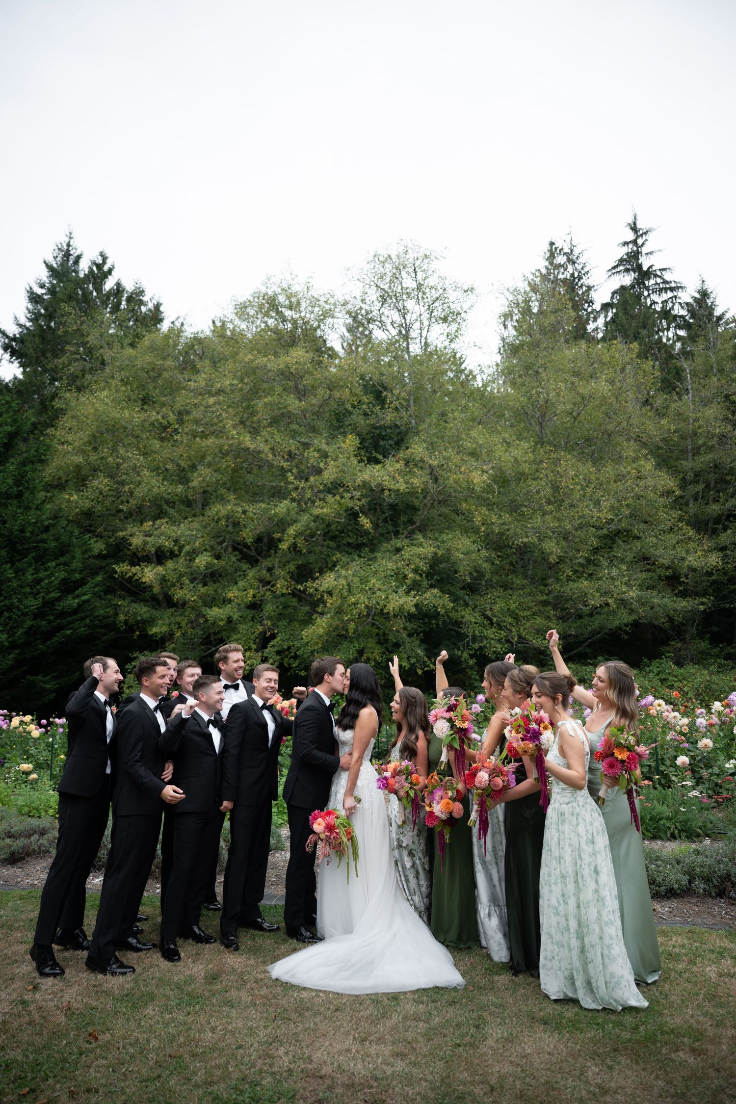 Wedding party celebrating outdoors with groomsmen in black tuxedos and bridesmaids in green dresses among flowers.