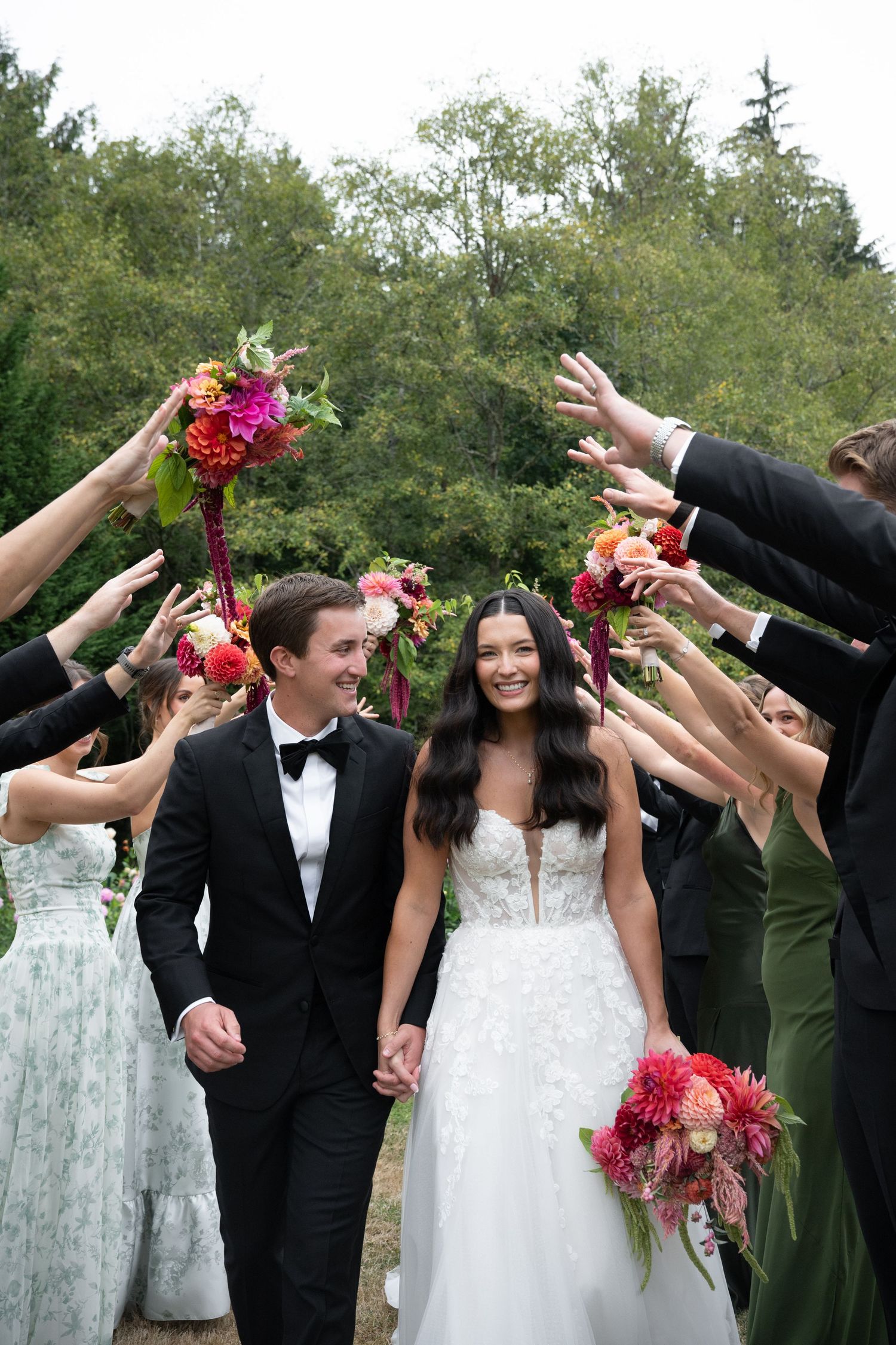 Bride and groom walk through floral arch tunnel created by wedding party holding colorful bouquets outdoors.