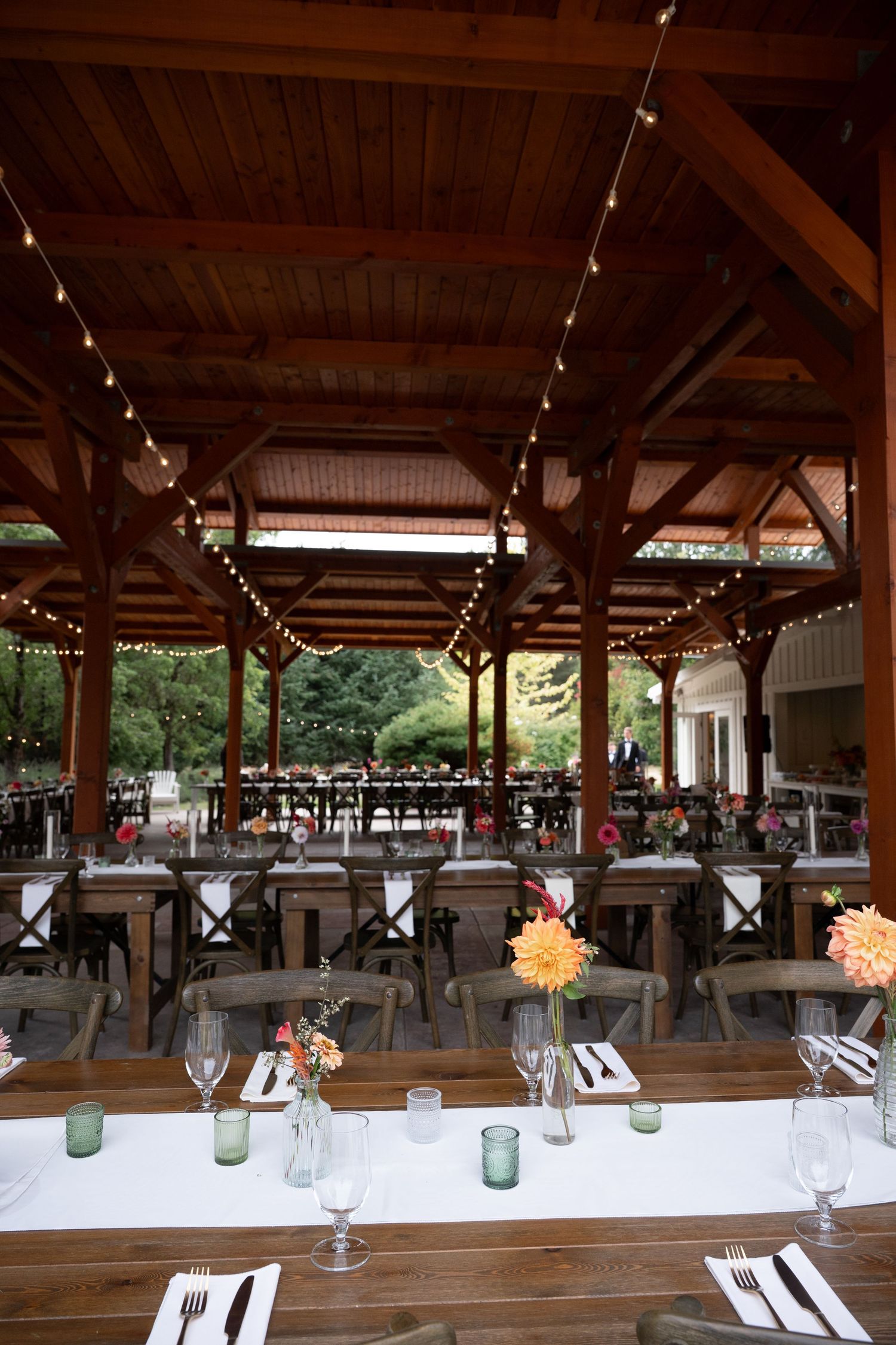 Elegant outdoor wedding reception pavilion with string lights, wooden tables, white linens, and colorful floral centerpieces.