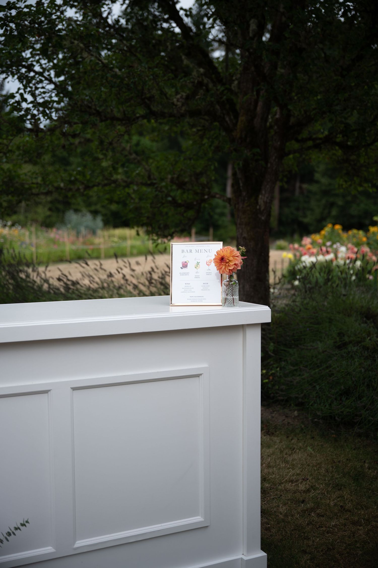 White outdoor bar with orange flower and menu card at garden wedding venue.