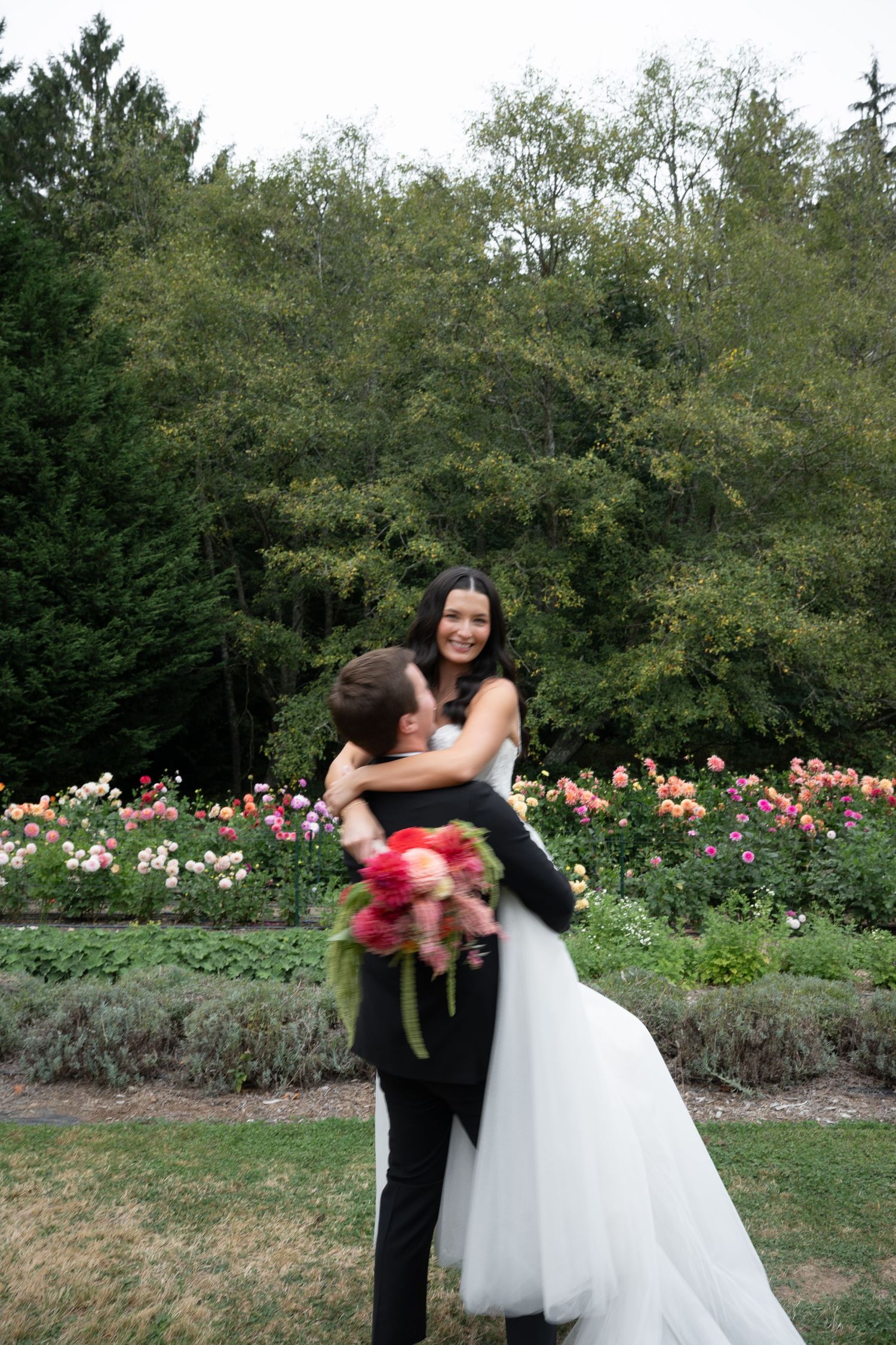 Bride and groom embrace in a lush garden wedding setting with colorful blooming roses and tall green trees in the background.