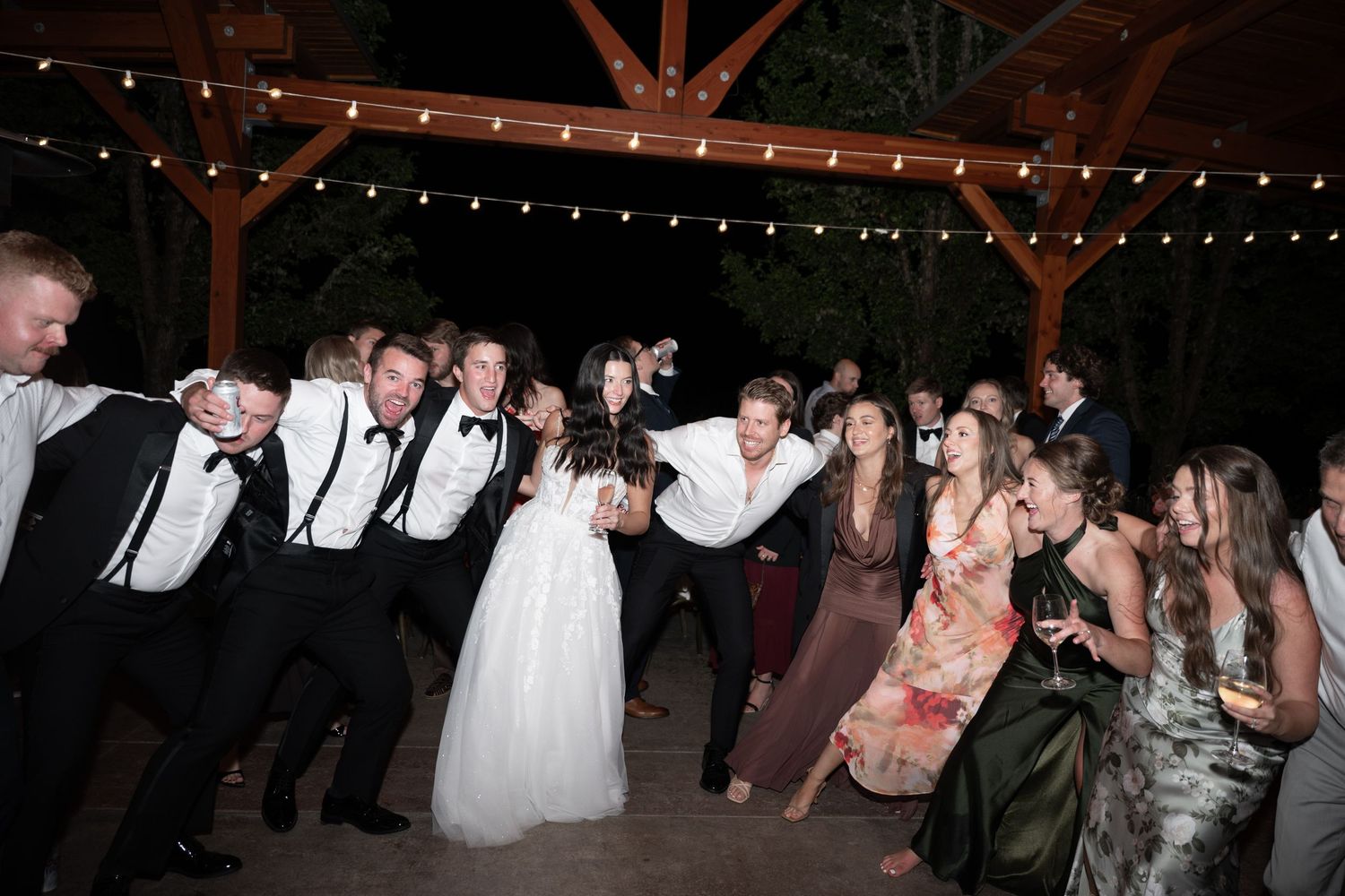 Wedding reception dance floor with bride, groom, and guests celebrating under string lights at an outdoor venue.