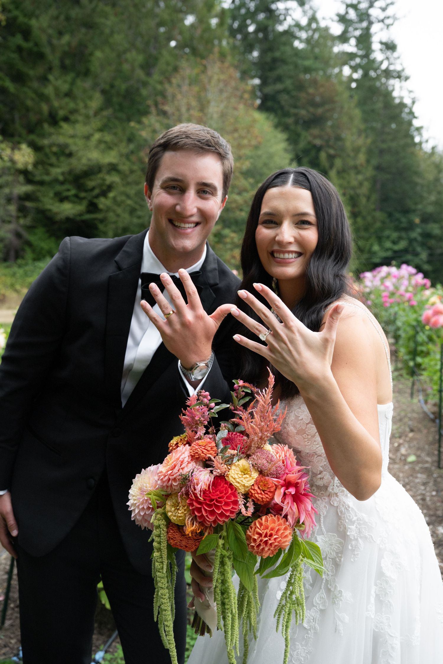 Smiling newlyweds showing off wedding rings while holding a vibrant bouquet of dahlias and wildflowers outdoors.