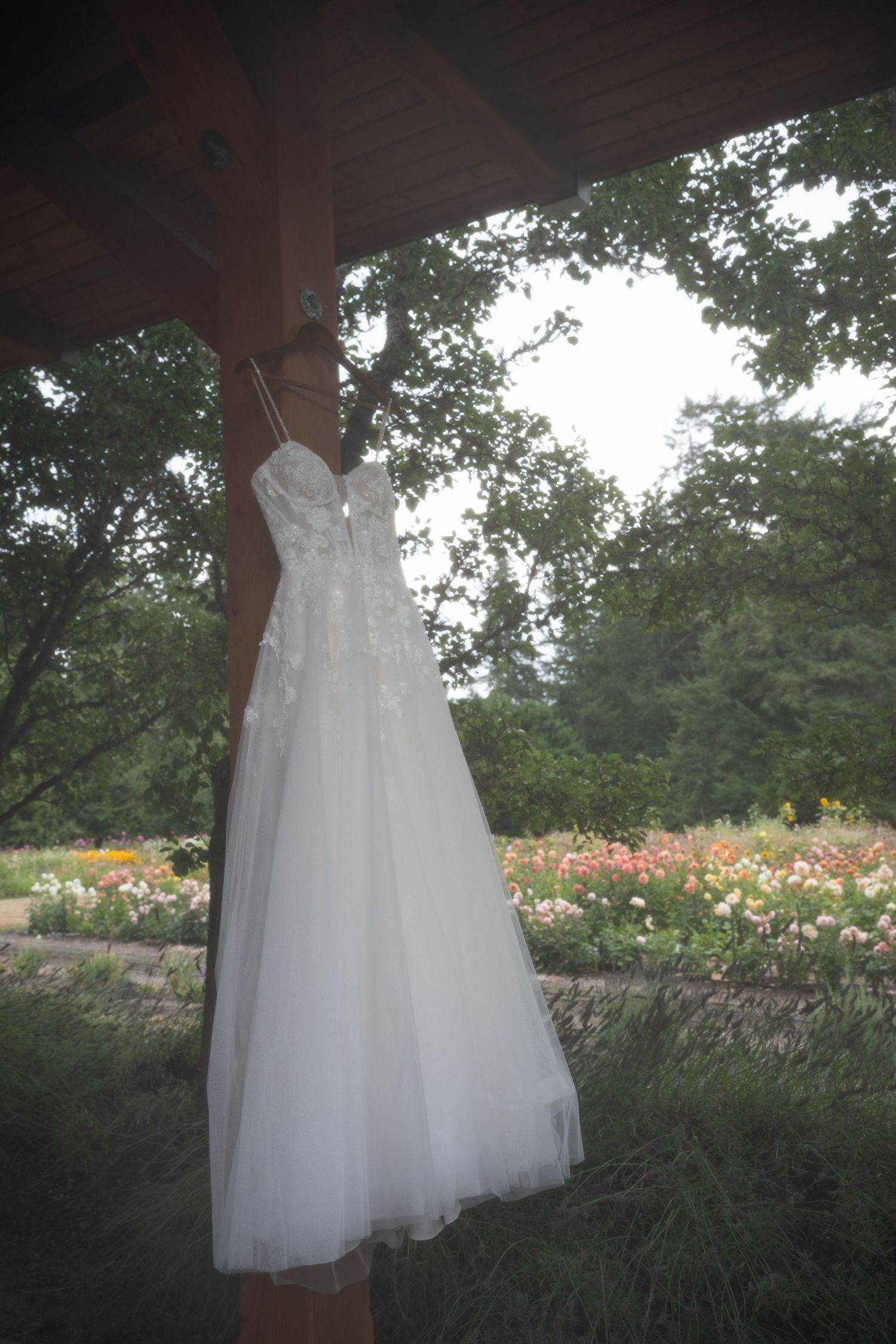 White lace wedding dress hanging on wooden post with colorful garden and green trees in background.