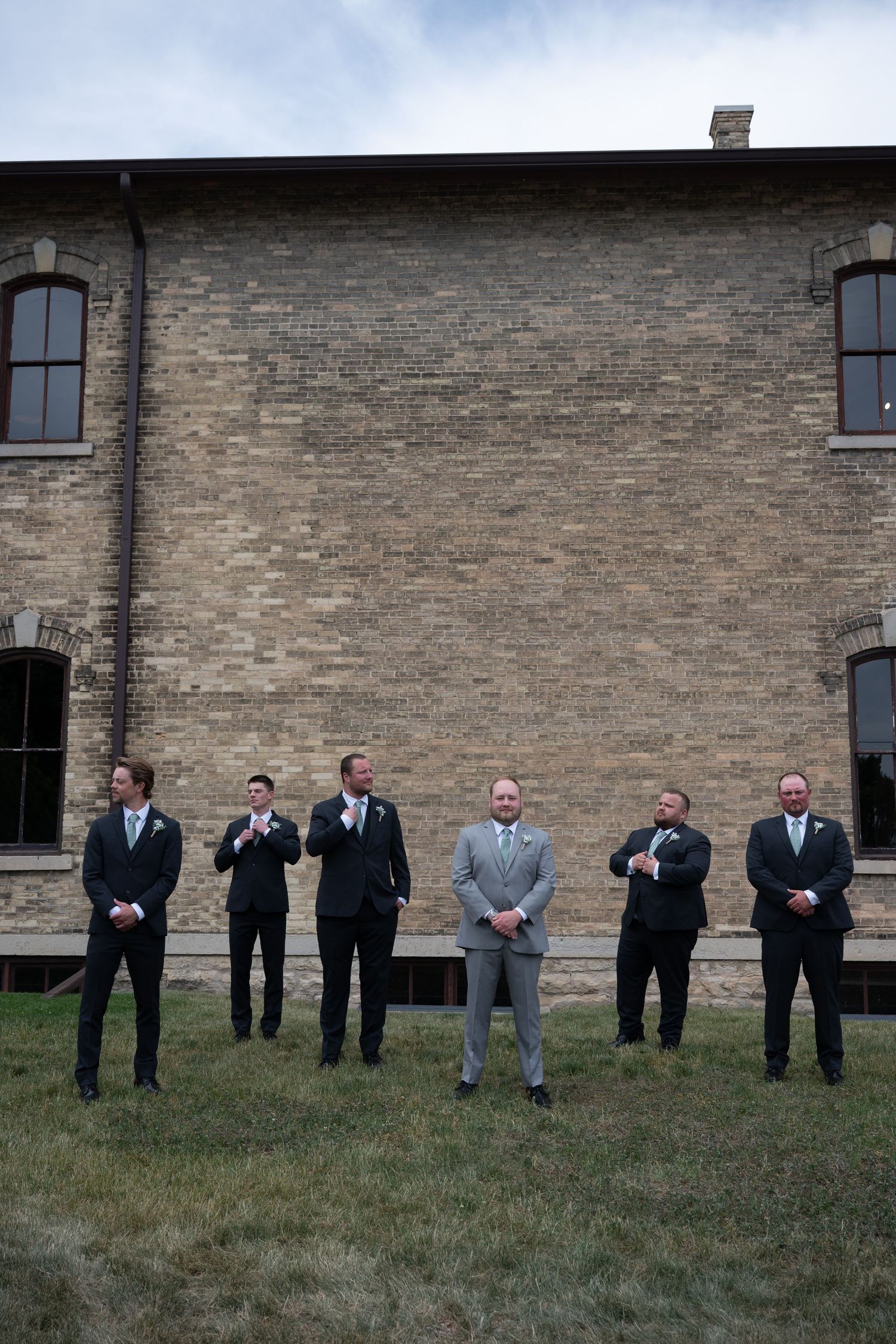 Groom in gray suit stands with five groomsmen in black suits outside historic brick building.