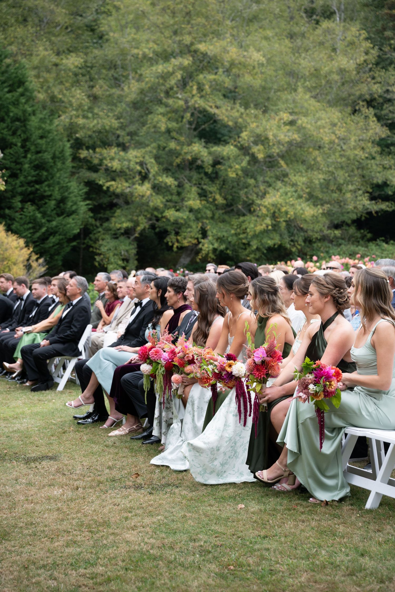 Bridesmaids in green dresses holding colorful bouquets seated at an outdoor wedding ceremony.