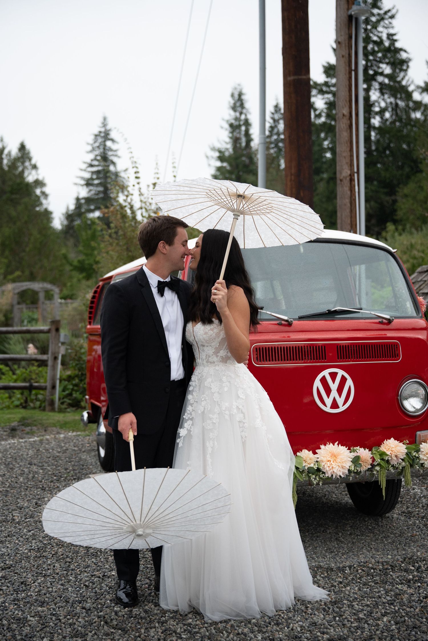 Bride and groom kiss behind white parasols in front of vintage red Volkswagen van.