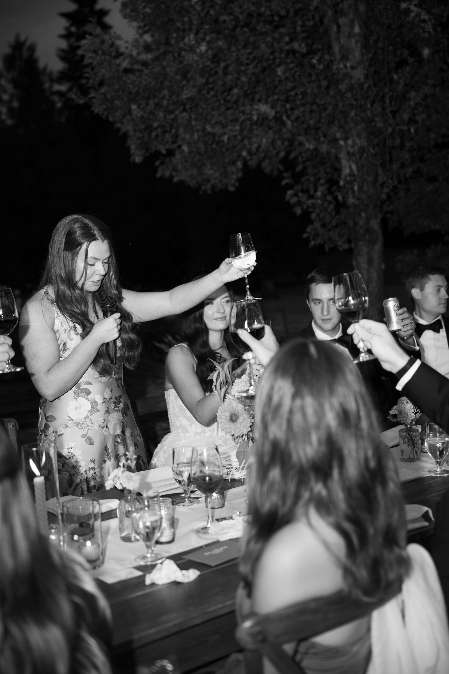 Wedding guests raise glasses in a toast at outdoor evening reception in black and white photo.