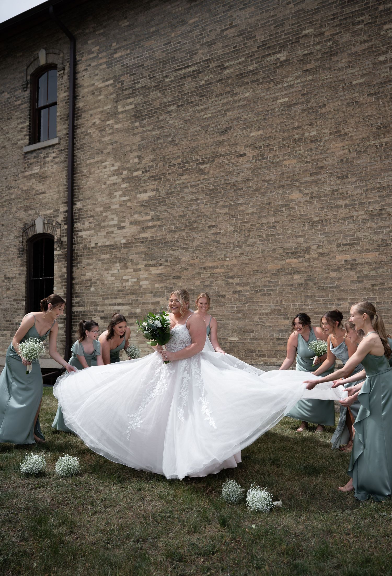 Bridesmaids in sage green dresses fluffing bride's white ballgown outside rustic brick building.