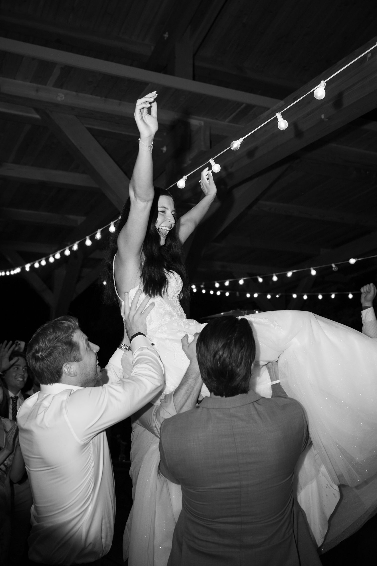 Bride lifted joyfully by groomsmen on dance floor with string lights at wedding reception, black and white.