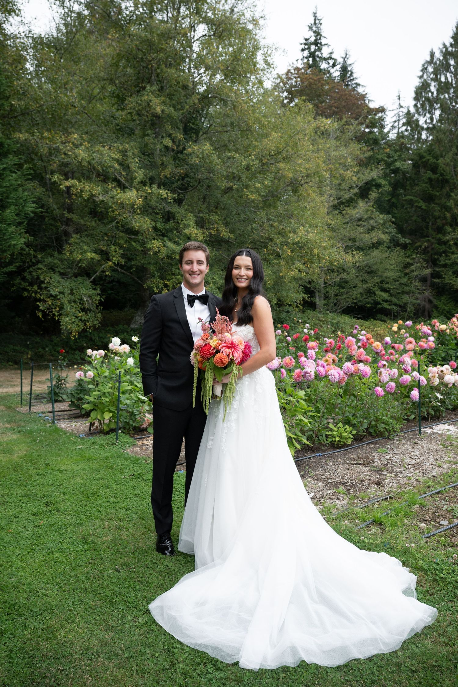 Smiling bride and groom pose together in front of vibrant pink dahlia garden on wedding day.