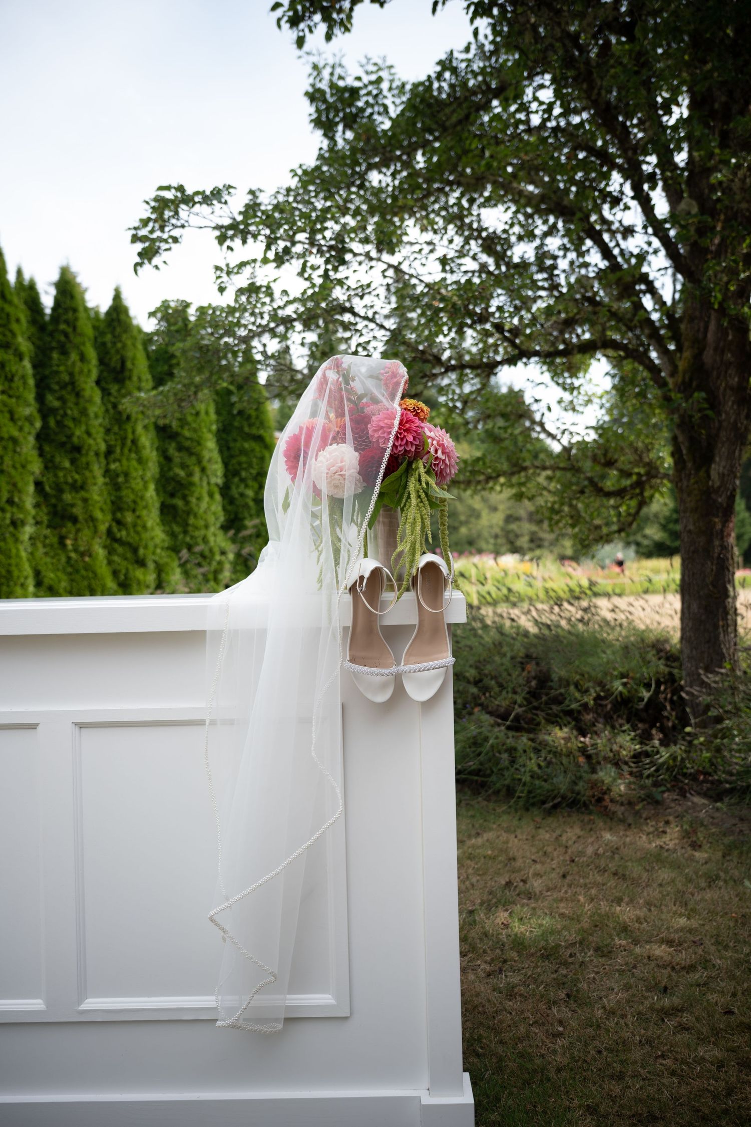 Bridal veil draped over white wedding bouquet and heels on white fence outdoors.