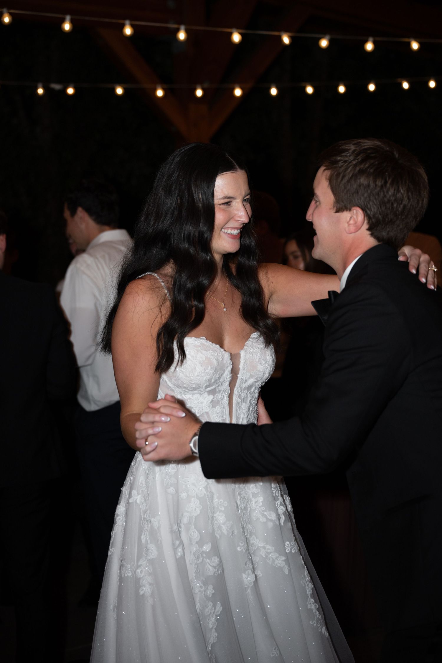 Bride in lace gown smiling while dancing with groom at wedding reception under string lights.