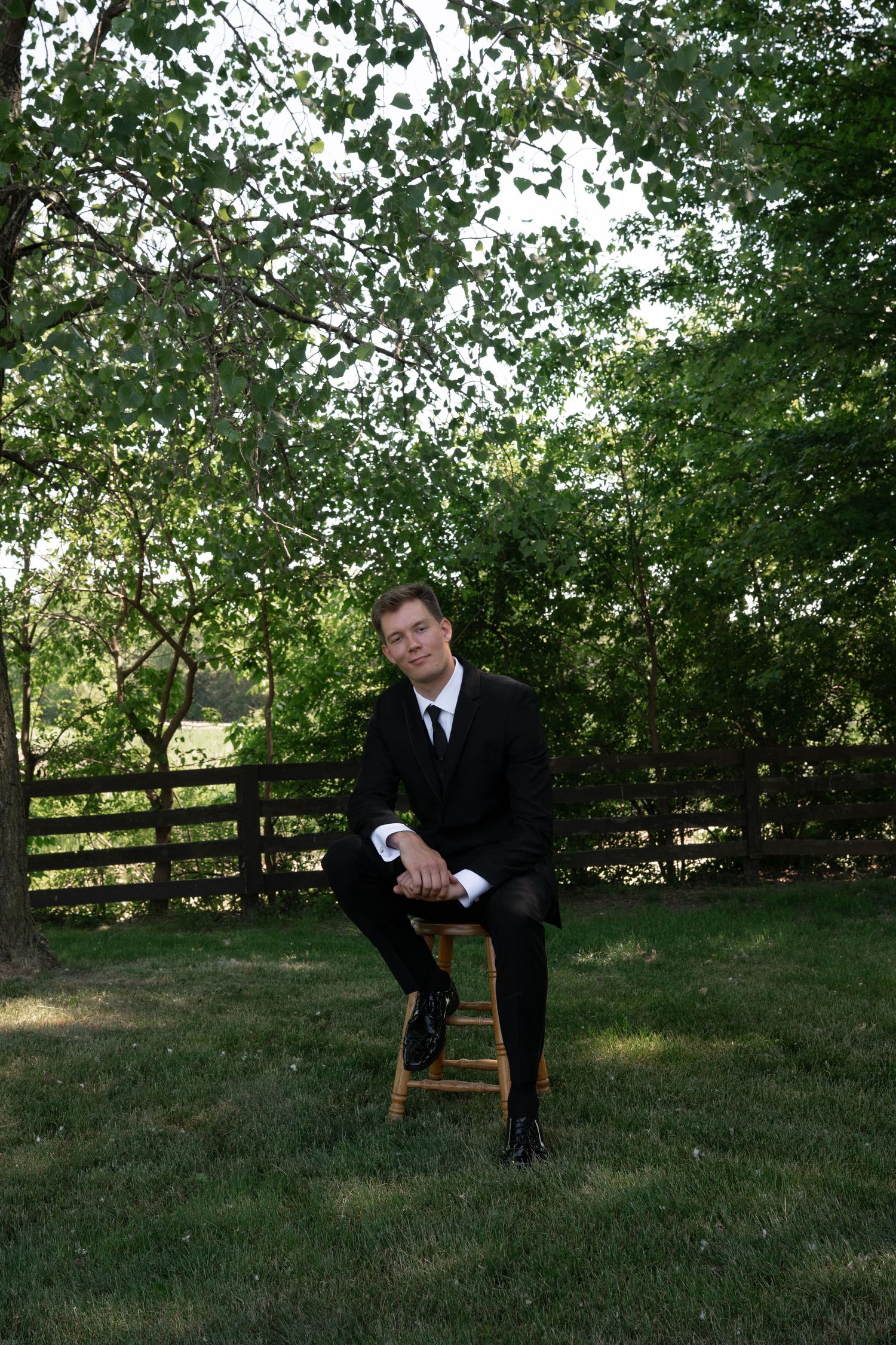 Elegant groom in black suit sitting on wooden stool in lush garden with wooden fence backdrop.