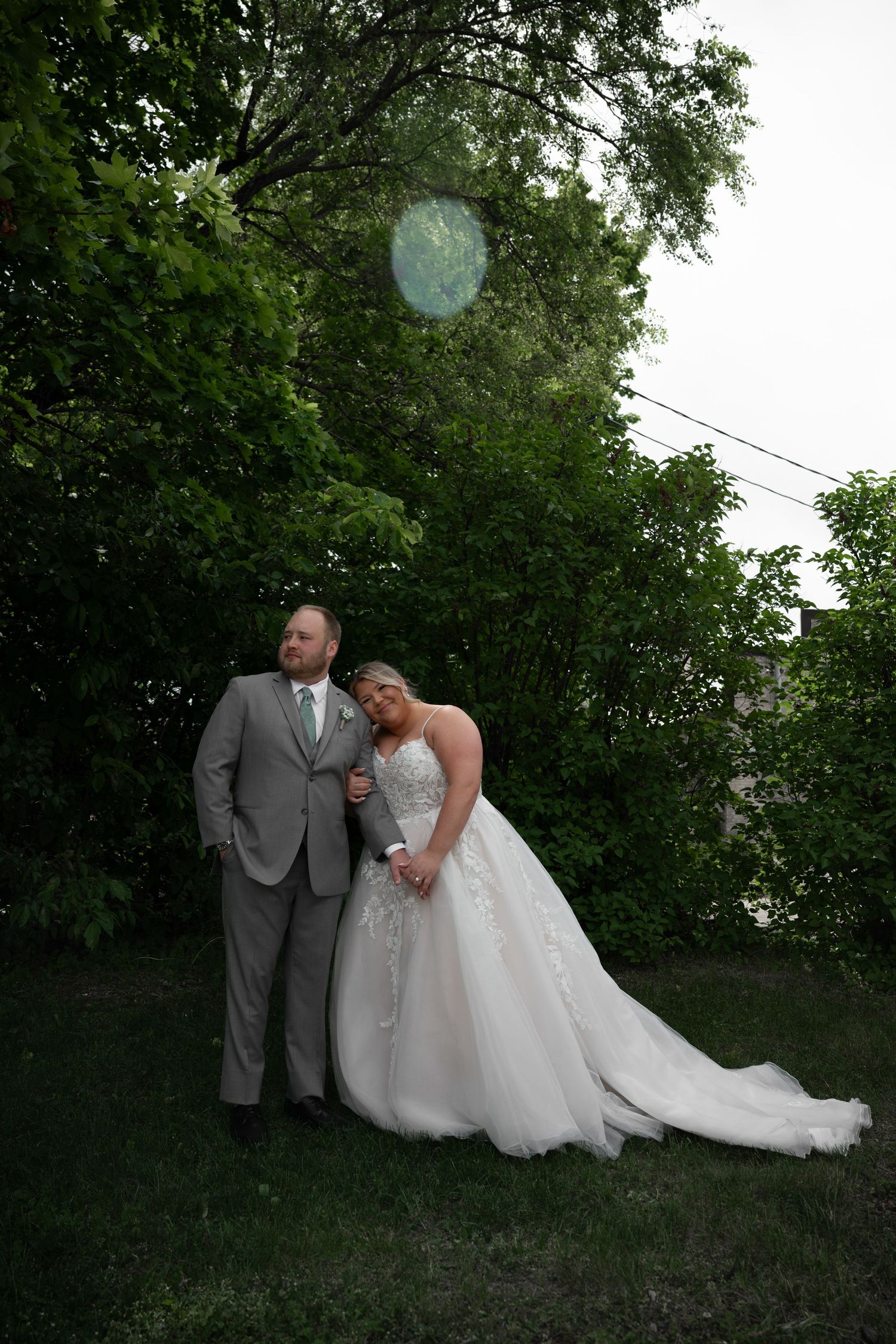 Bride and groom pose together under lush green trees with lens flare in evening light.