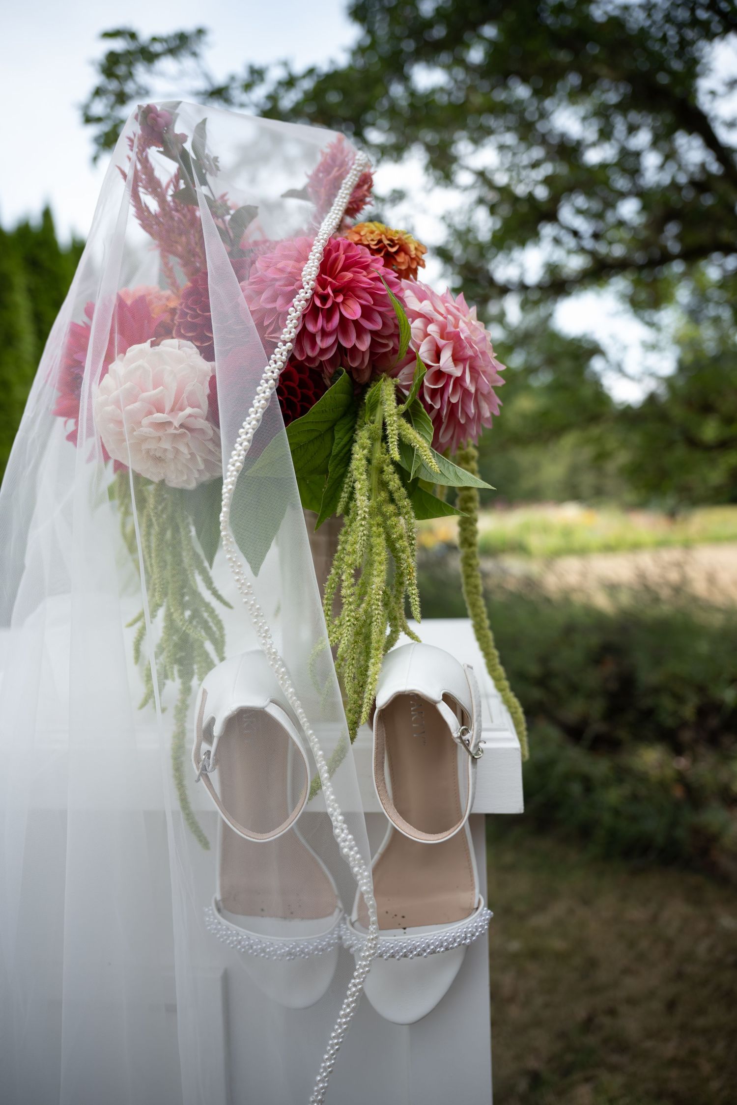 White bridal heels and veil displayed beside a vibrant pink dahlia wedding bouquet outdoors.