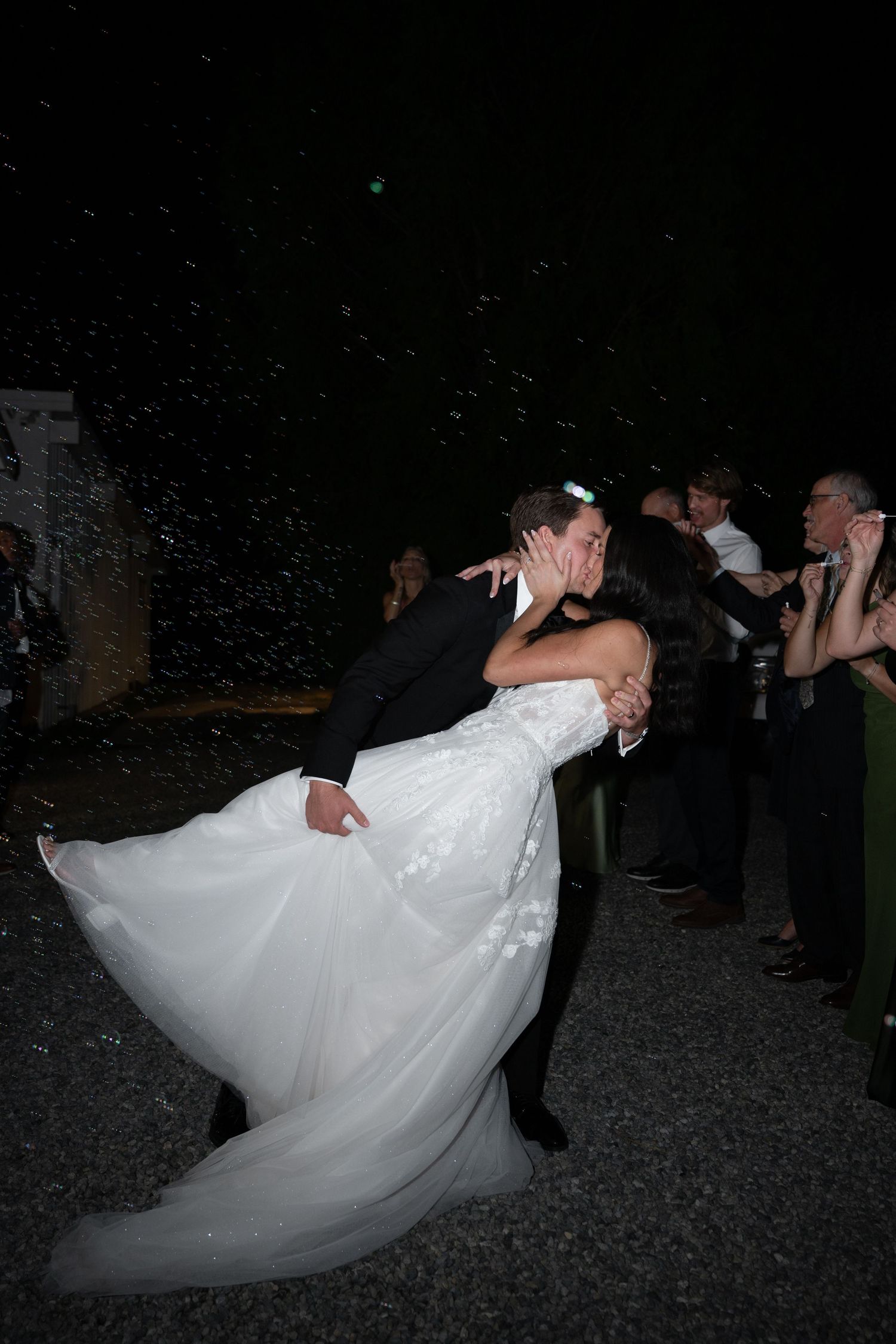 Groom dips bride for a romantic kiss during outdoor wedding reception at night with guests watching.