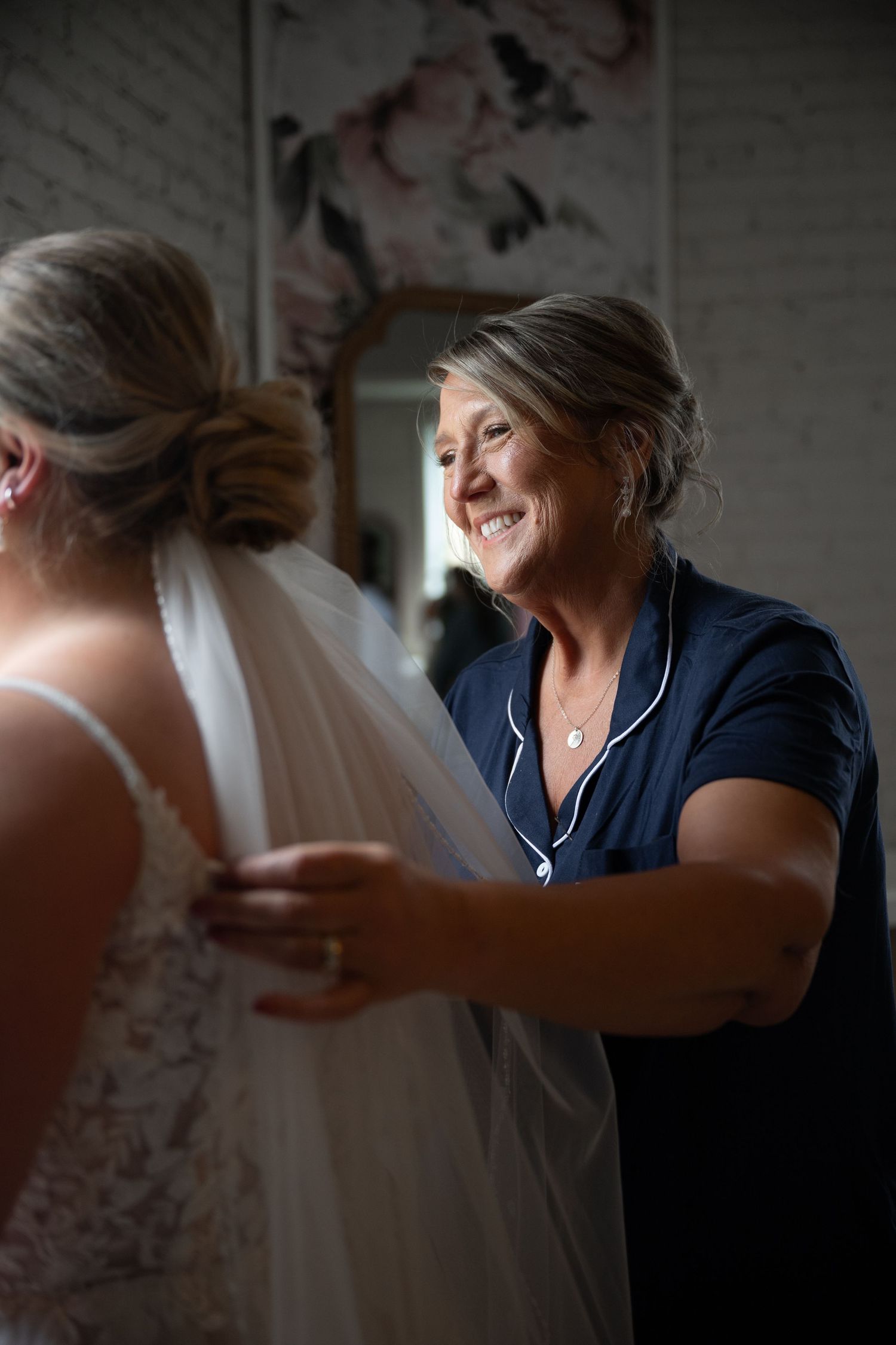 Smiling woman in navy pajamas helping bride with veil in bright dressing room with mirror.