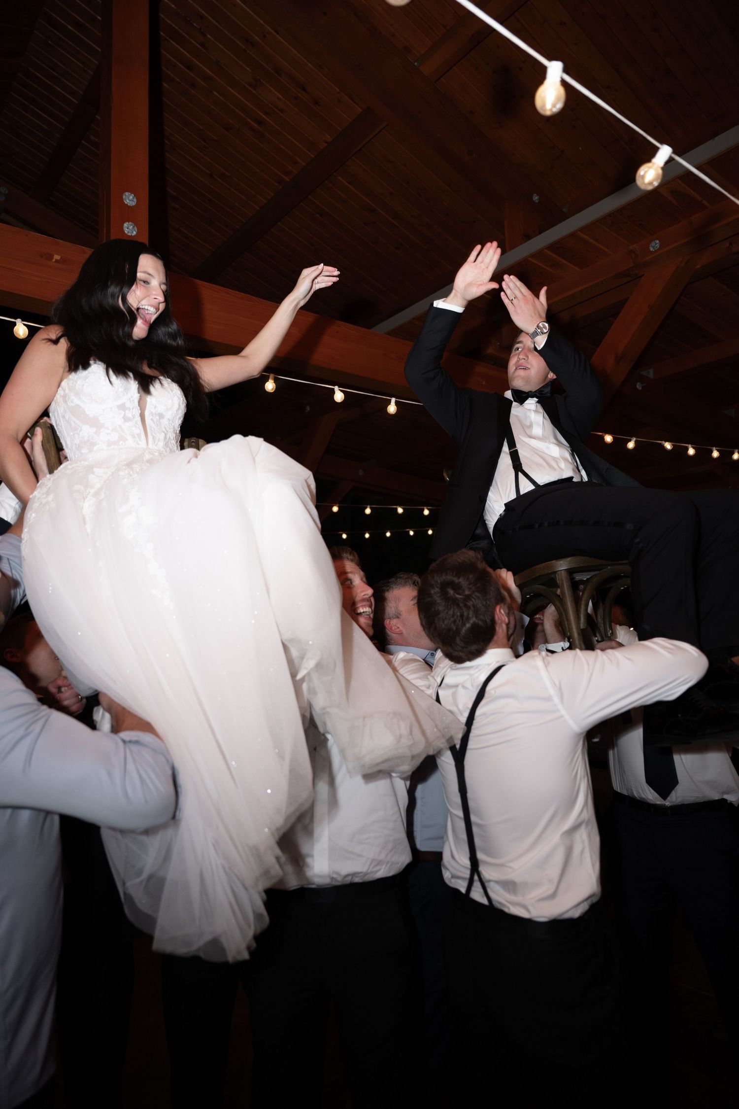 Bride and groom lifted by guests during lively wedding reception dance celebration in rustic venue with string lights.