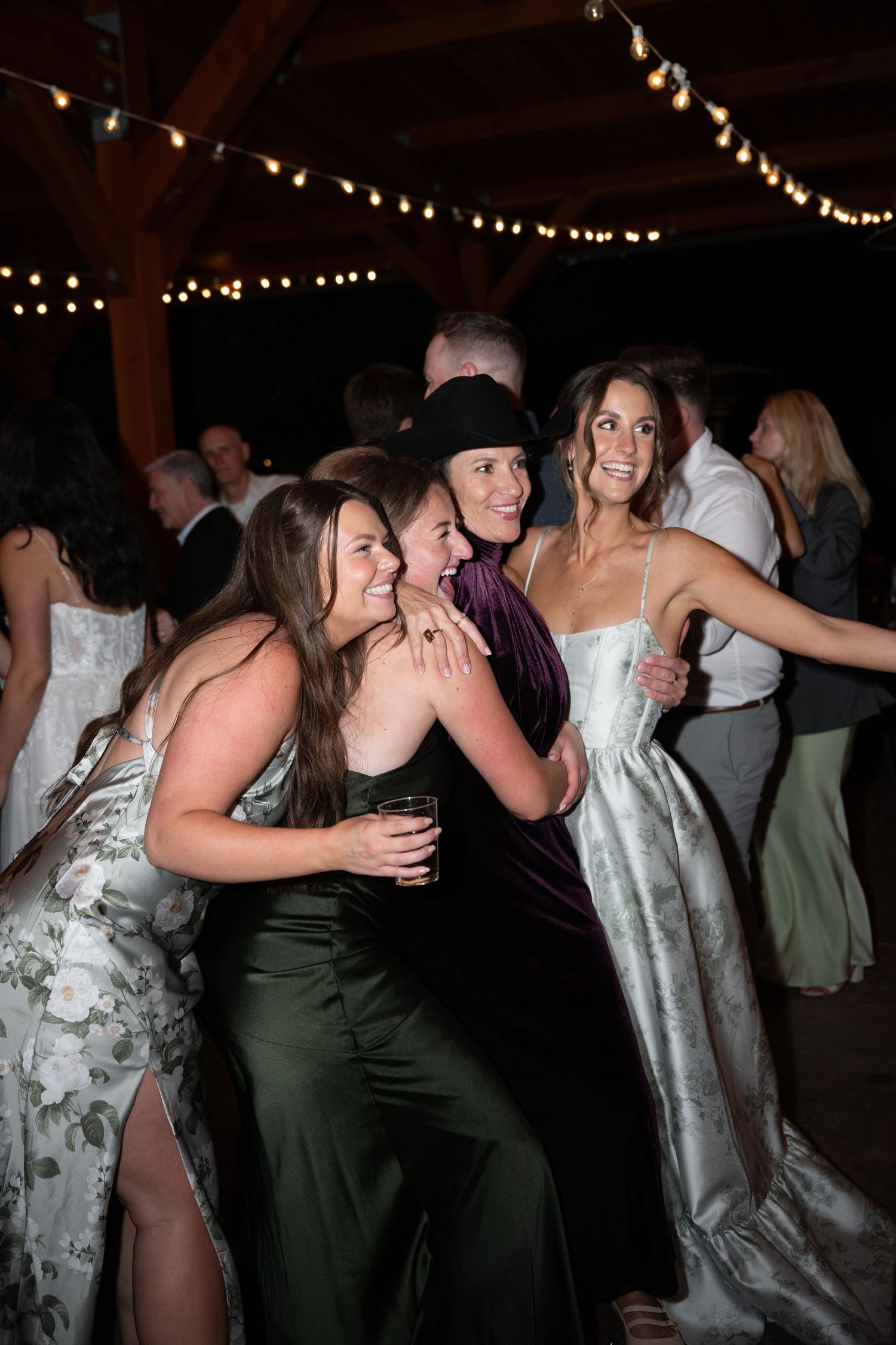 Bridesmaids and wedding guests dancing joyfully under string lights at lively indoor wedding reception at night.