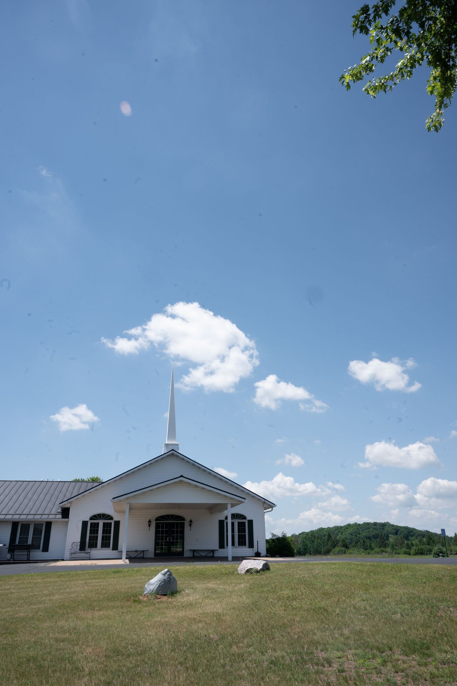 White church with tall steeple sits on green lawn under bright blue sky with scattered clouds and wooded hills behind.