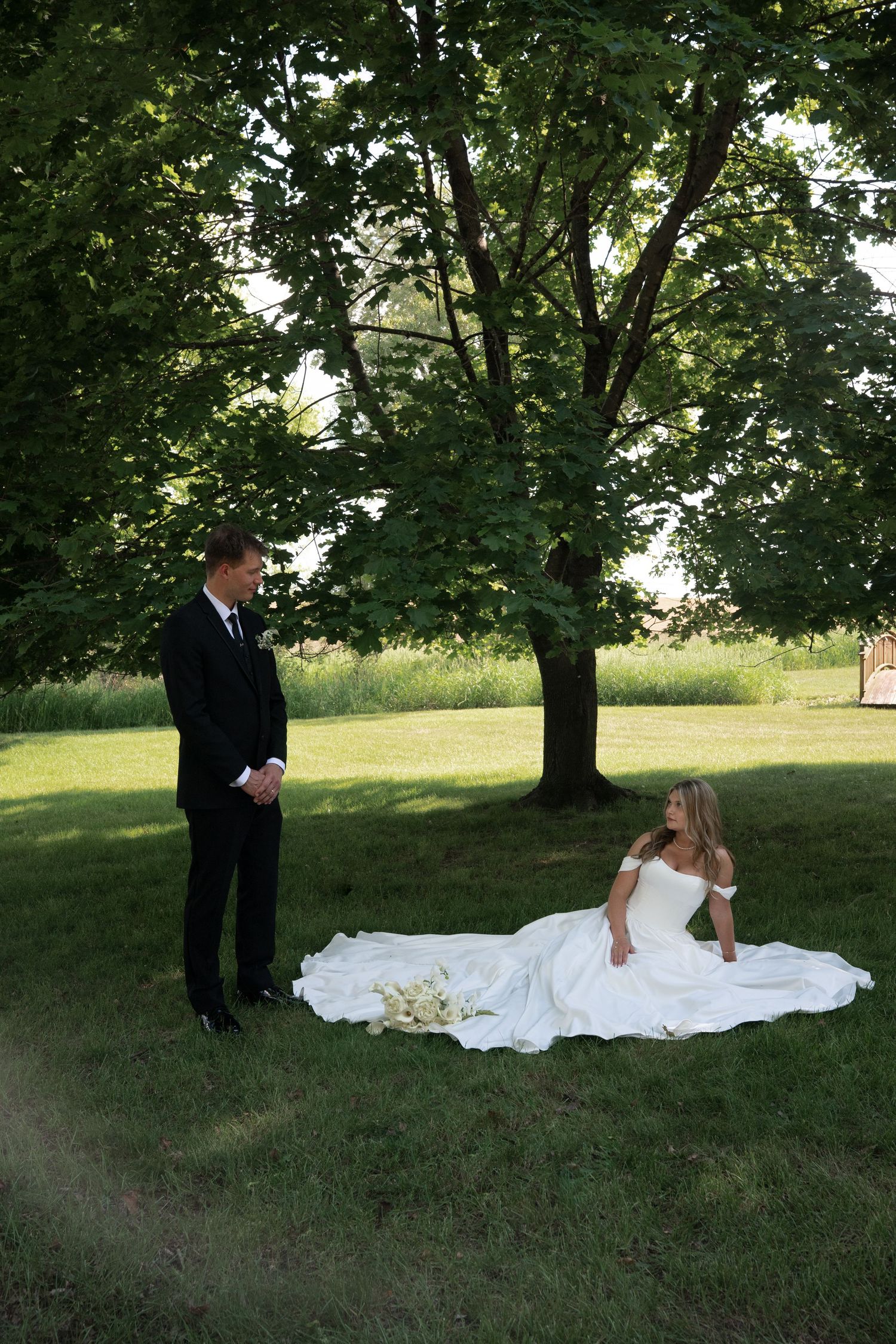 A bride in a white gown sits on green grass while her groom stands nearby under large trees at an outdoor wedding.