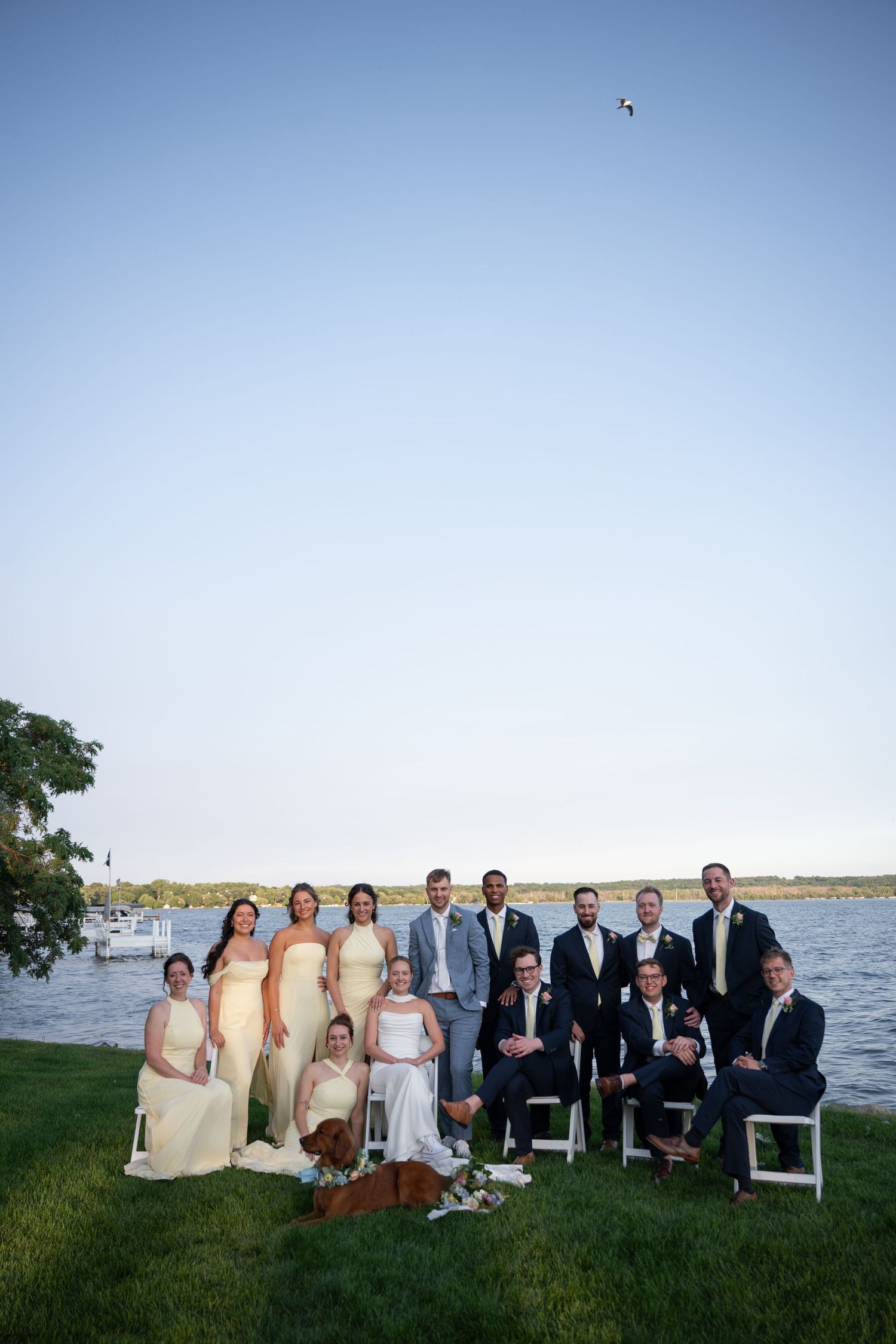 Wedding party in yellow and navy attire pose together on lakeside lawn at golden hour.