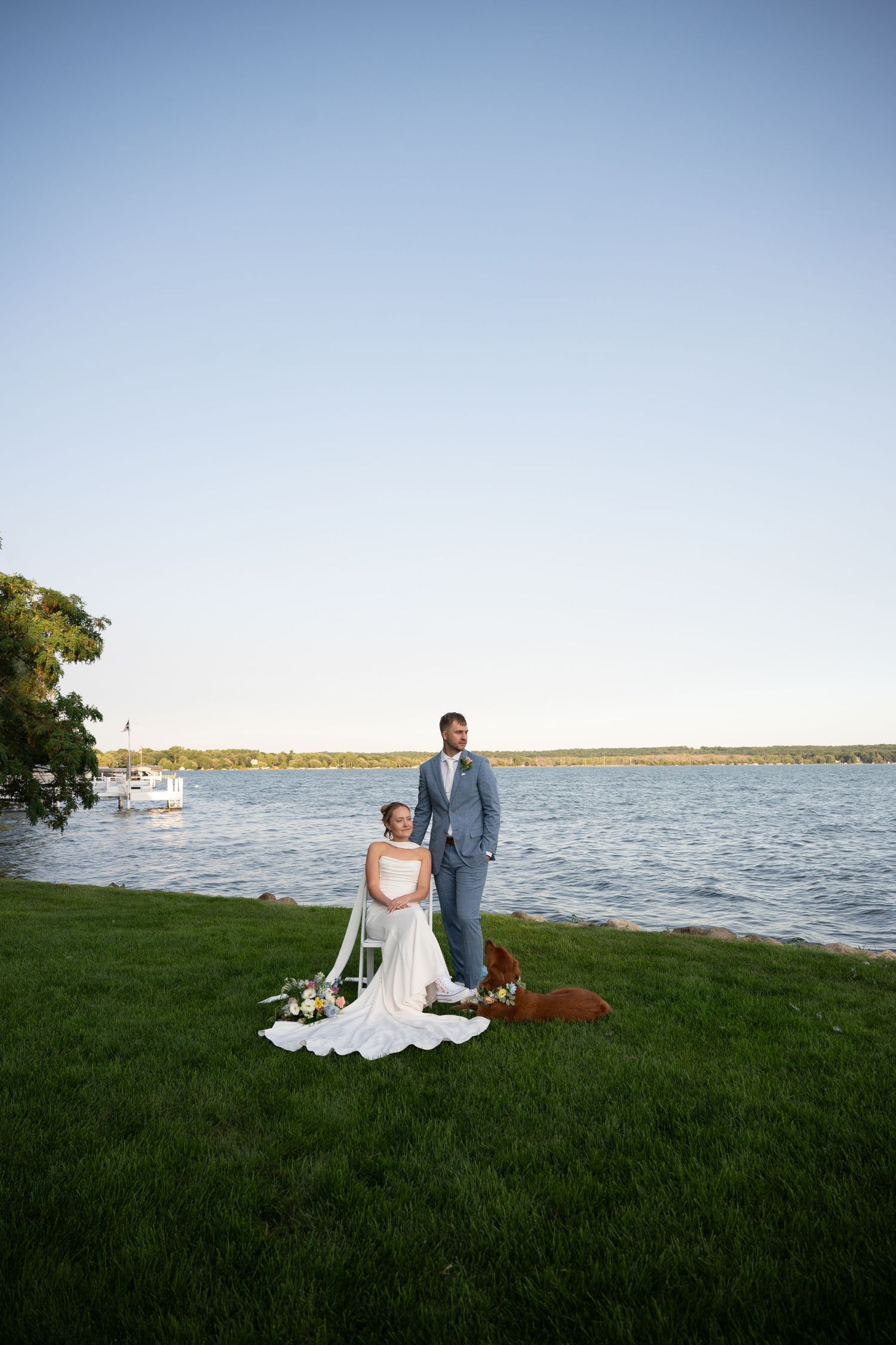 Bride in white gown and groom in gray suit pose by a serene lakeside on lush green grass during golden hour.