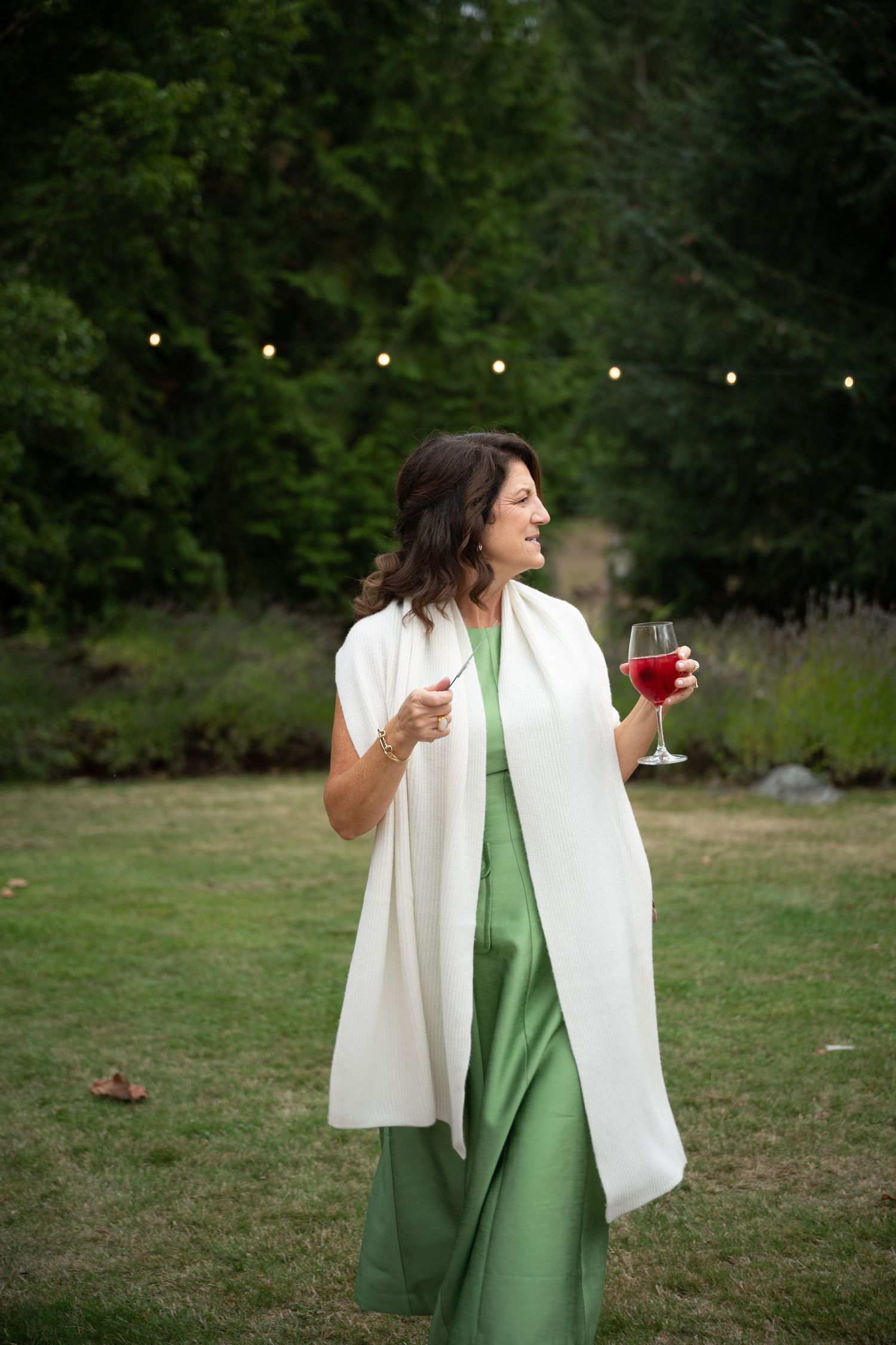 Elegant woman in green dress and white wrap holds red wine glass at outdoor evening wedding reception.