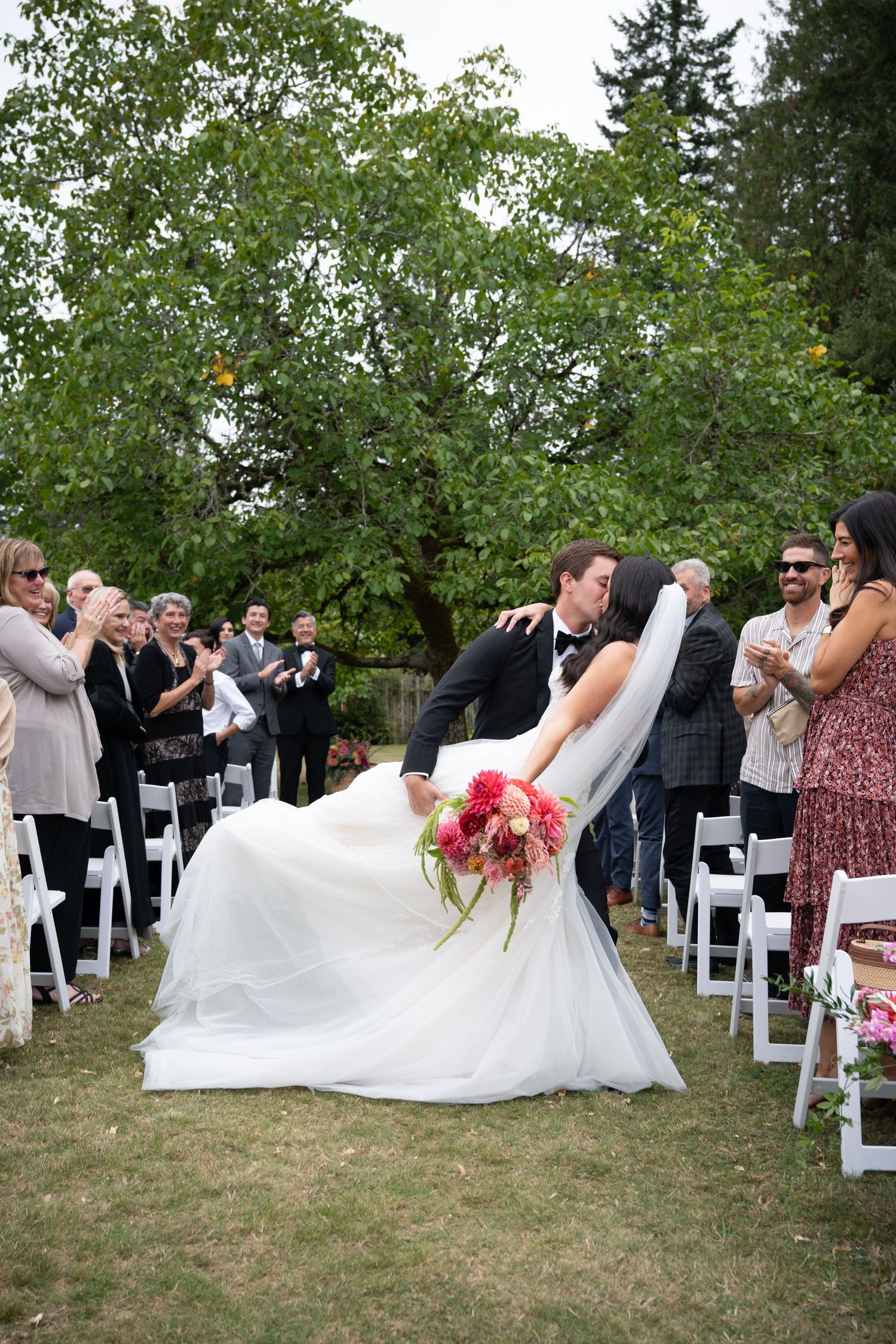 Groom dips bride for a kiss at outdoor wedding ceremony while guests cheer and applaud on a lush green lawn.