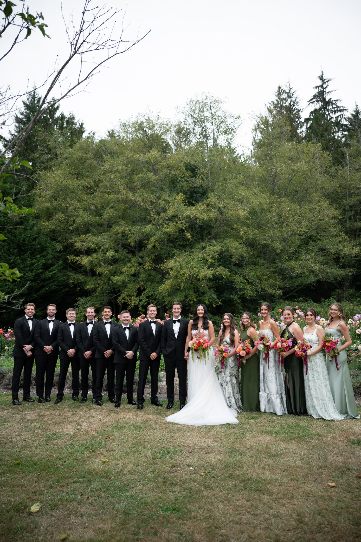 Wedding party with groomsmen in black tuxedos and bridesmaids in green dresses pose outdoors.