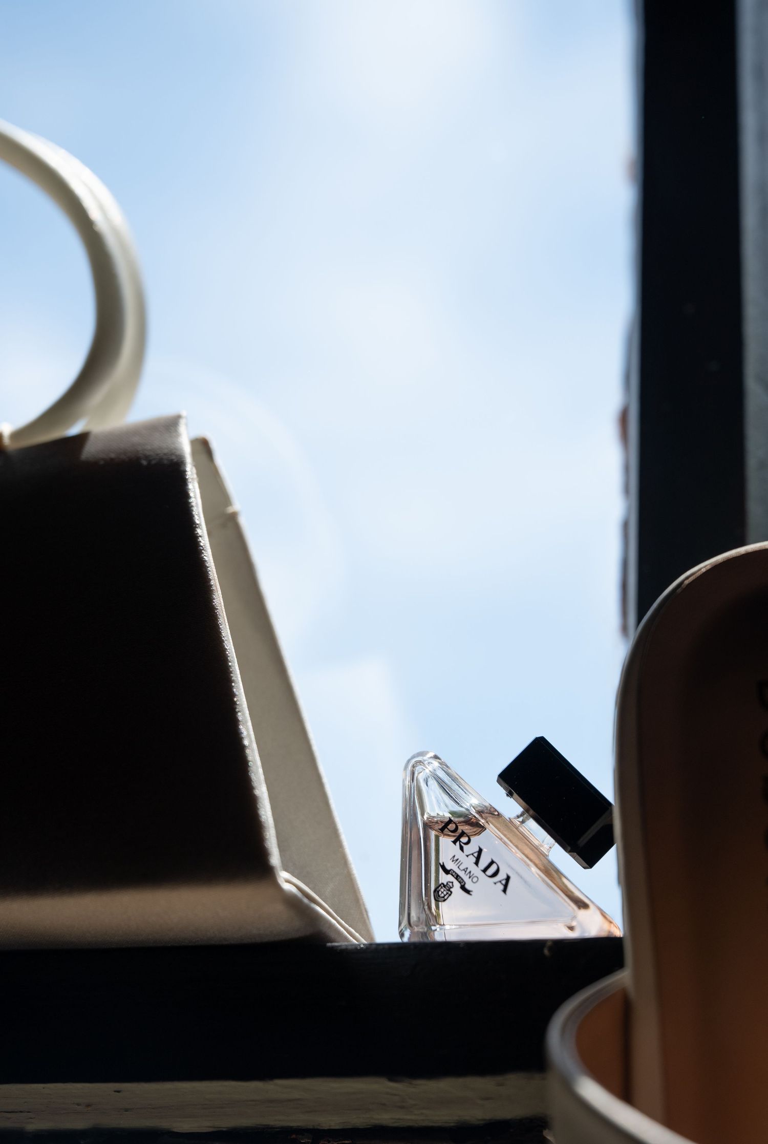 Miniature Prada perfume bottle on a dark surface with blue sky and white clouds in background.