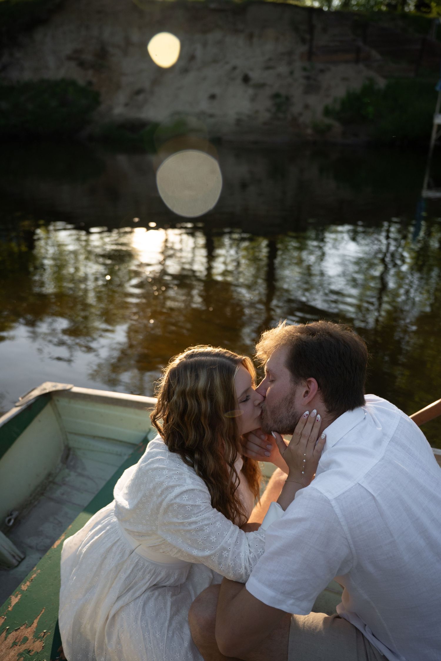 Romantic couple sharing a kiss in a rowboat on a tranquil river near a stone bridge at sunset.
